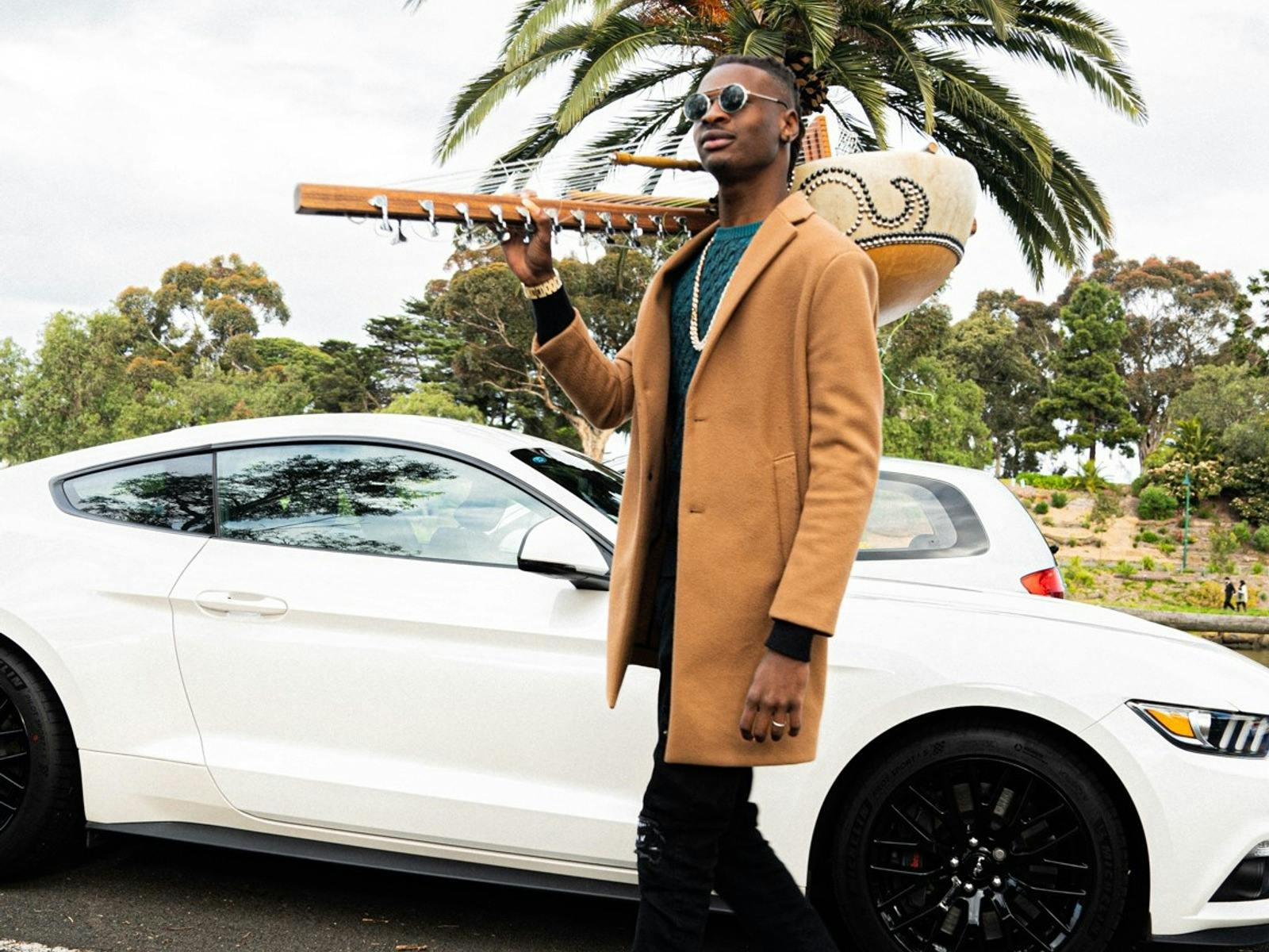 Amadou Suso holds his instrument, the kore, over his shoulder as he walks in front of a tree and car