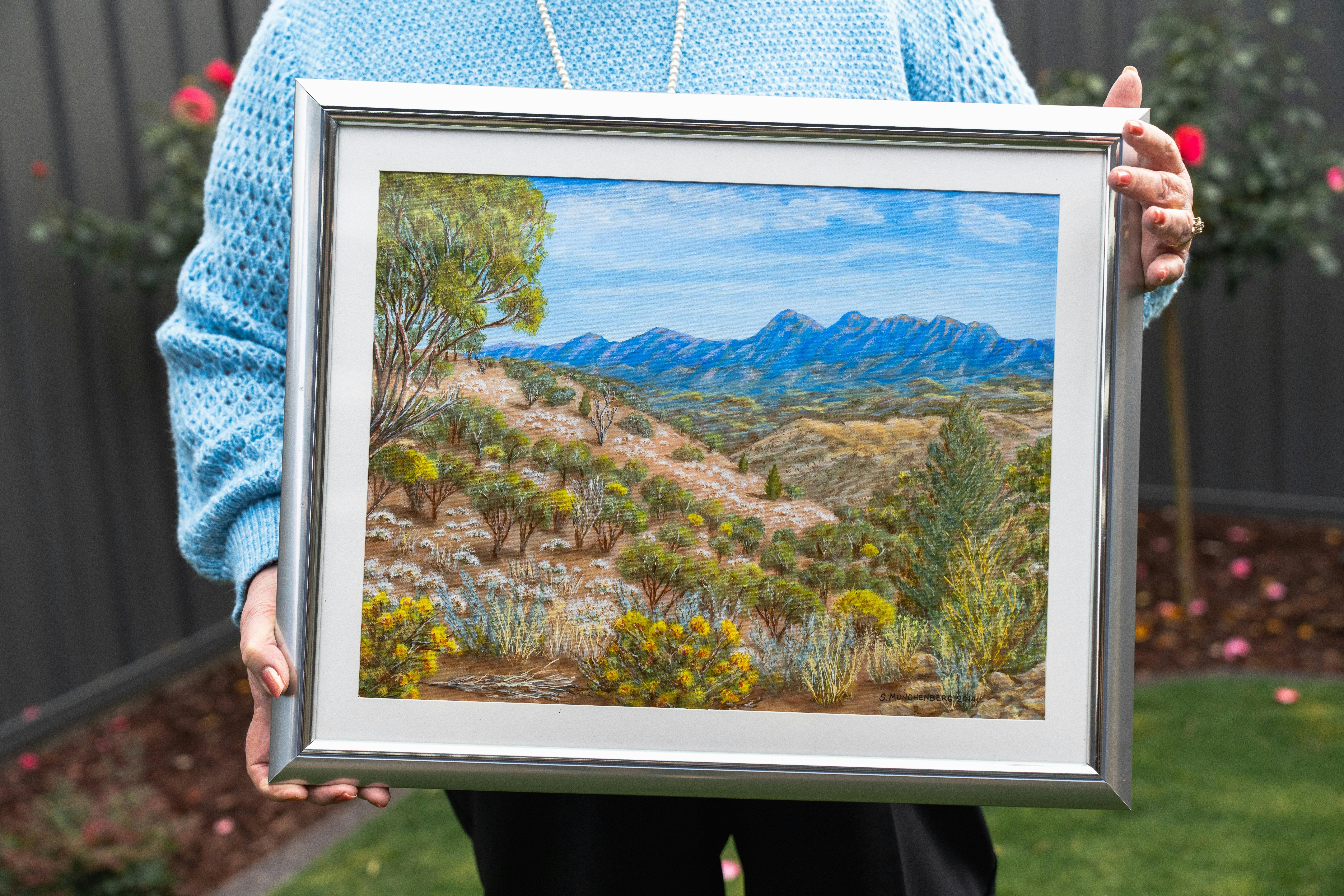 Older artist holding a framed painting of a rugged Australian mountain landscape.