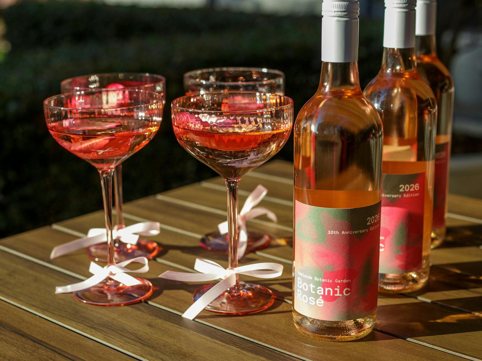Two ruby-pink rosé glasses with white ribbon bows sit beside three bottles of botanical rosé