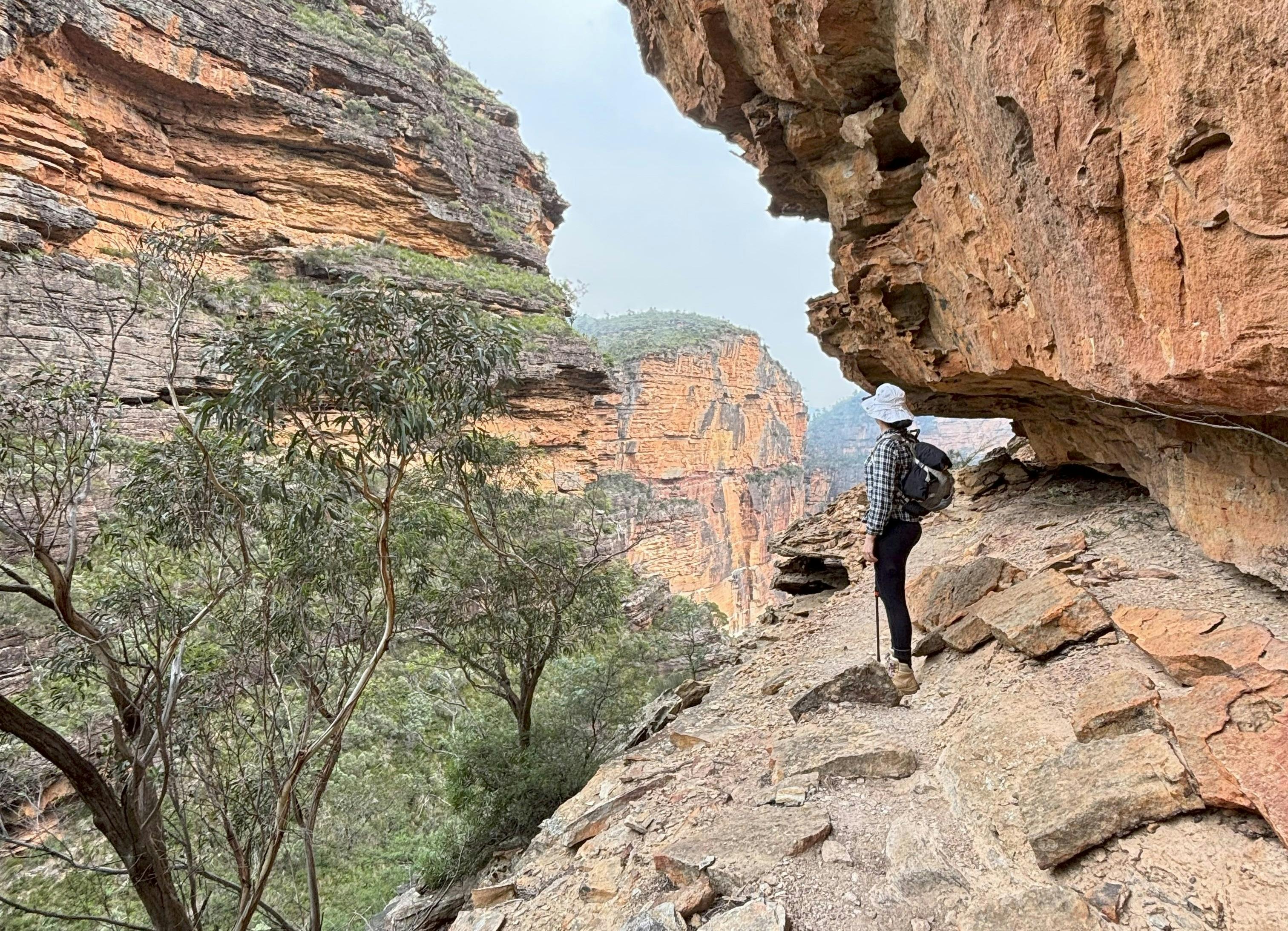 A woman wearing black leggings, checkered shirt and a backpack standing on the edge of a cliff.