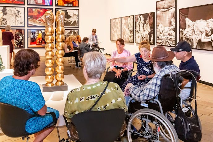 A group of people, some in wheelchairs, sit around a sculpture listening to a guide