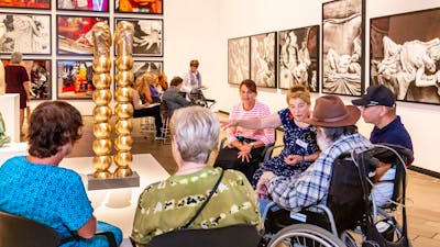A group of people, some in wheelchairs, sit around a sculpture listening to a guide
