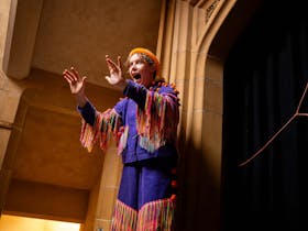 A lady standing in front of a door wearing a colourful costume with tassels and a hat