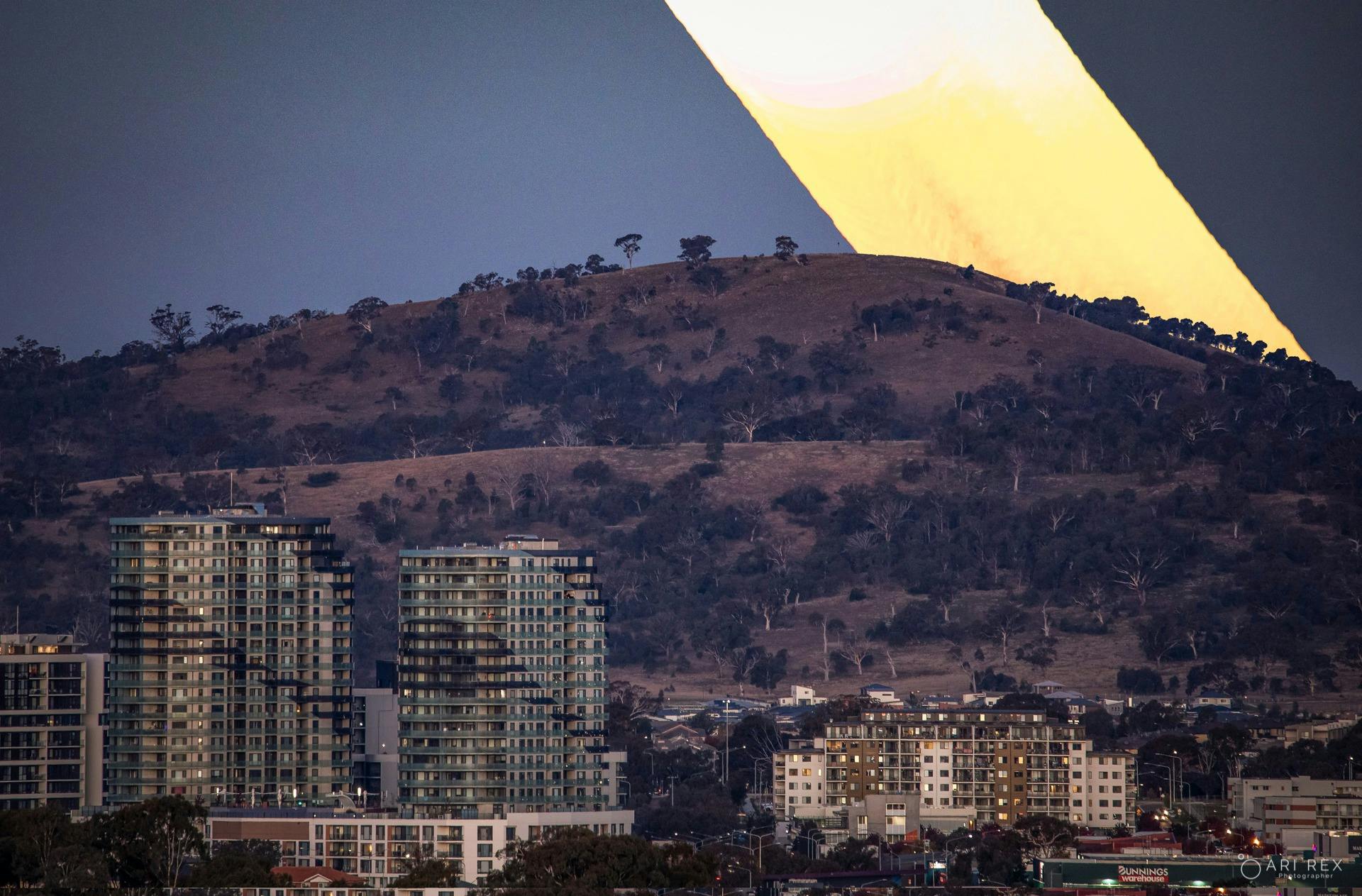 Last light falls across rugged hills above a town at dusk, beneath a deep blue sky with warm light n