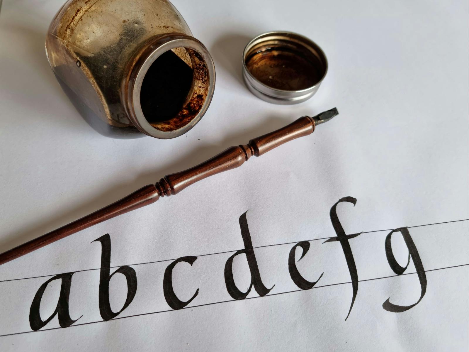 Handwritten letters ABCDEFG calligraphy on white paper with ink well and pen nearby