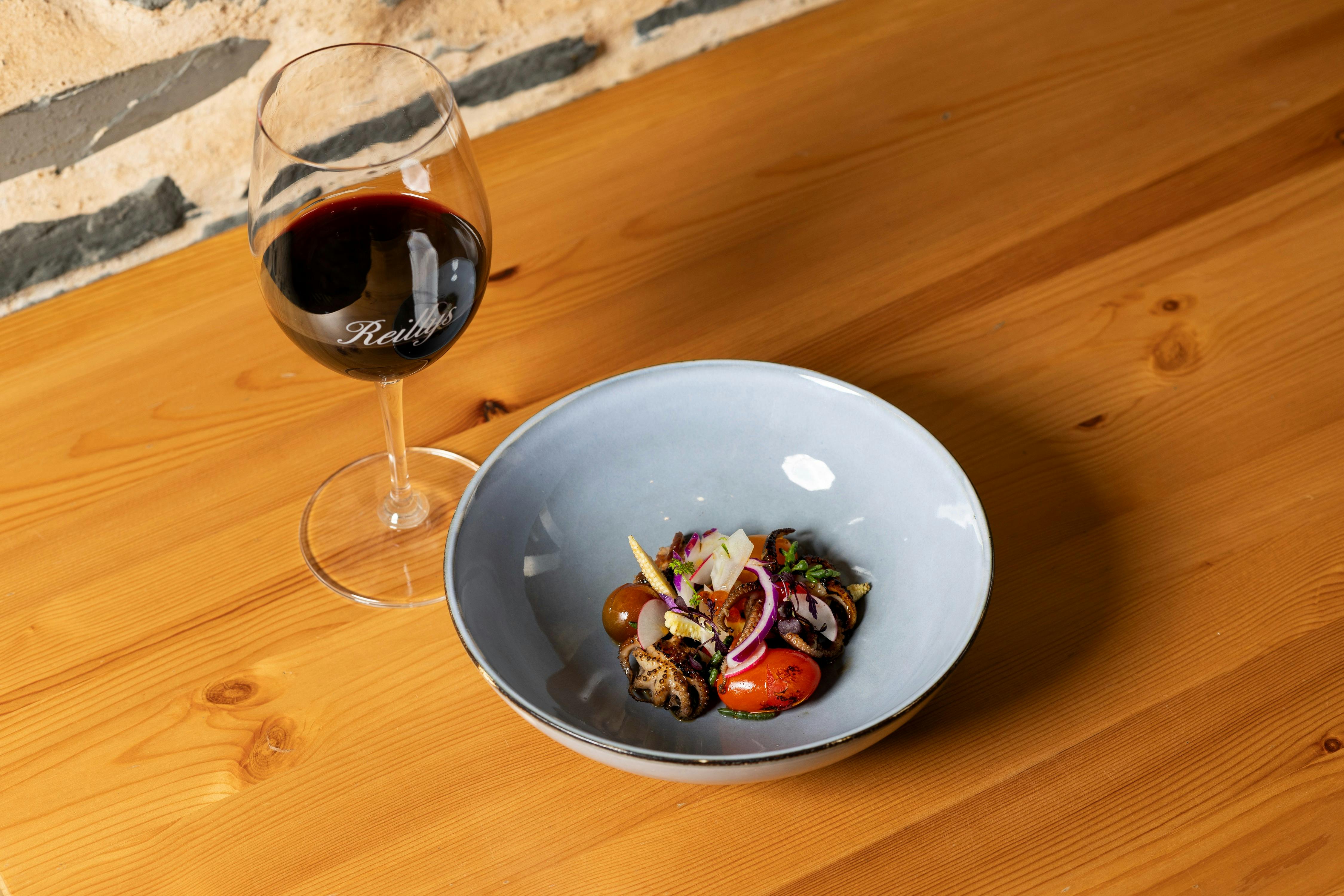 Bowl of fresh grilled baby octopus salad next to glass of red wine on a wooden table