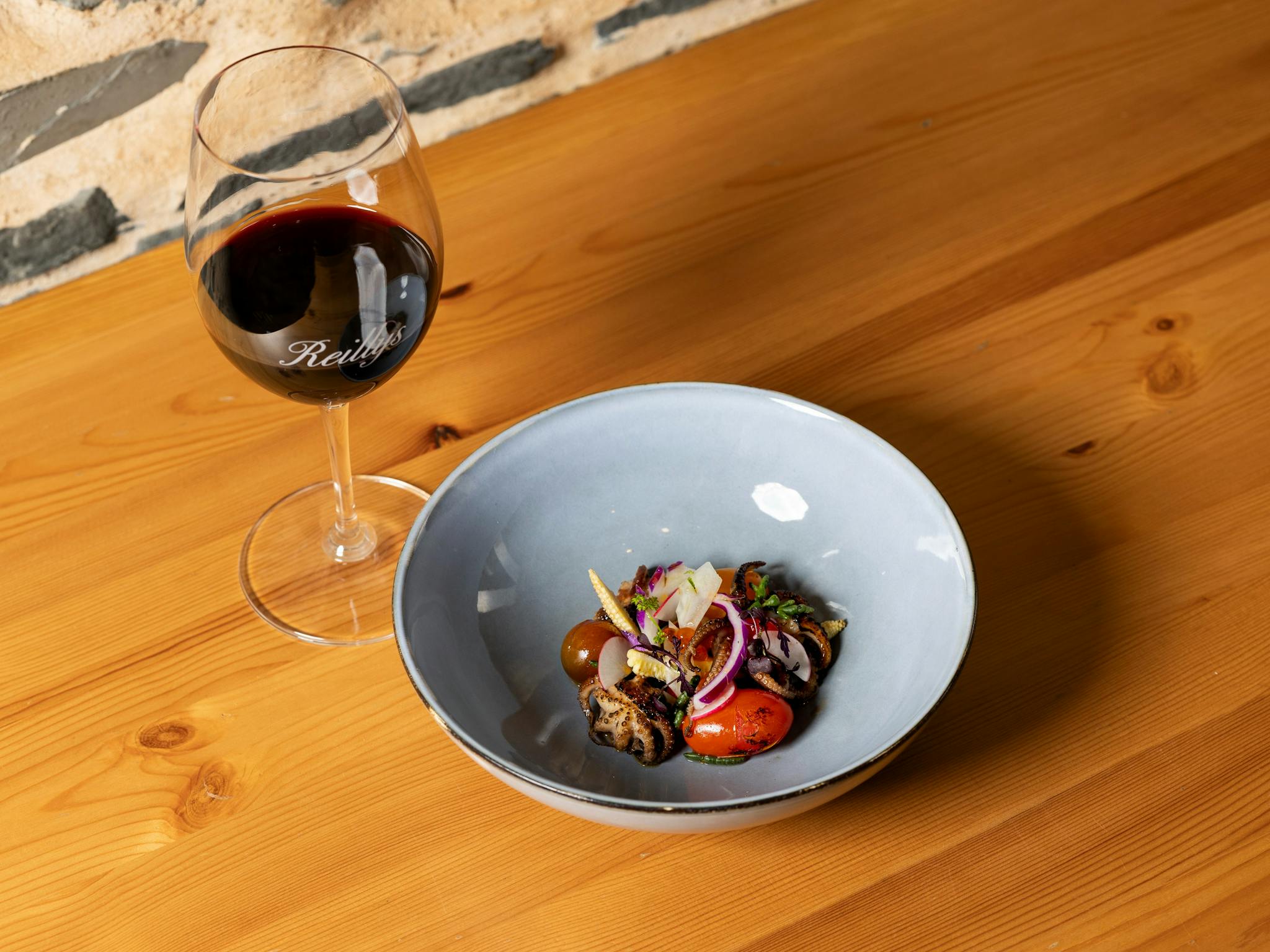 Bowl of fresh grilled baby octopus salad next to glass of red wine on a wooden table