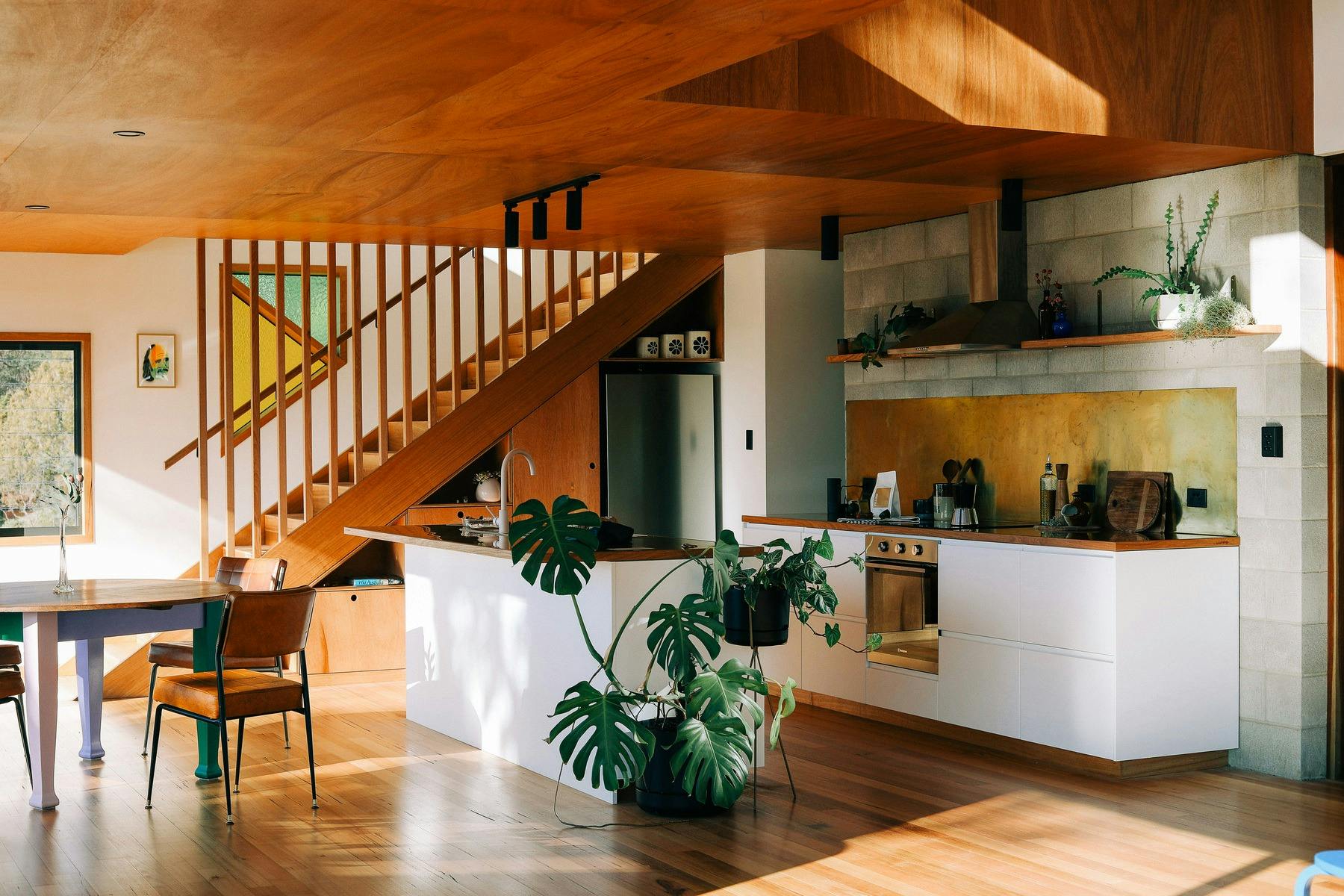 A kitchen with brass spashblack and large monstera plant with a timber staircase behind and table.
