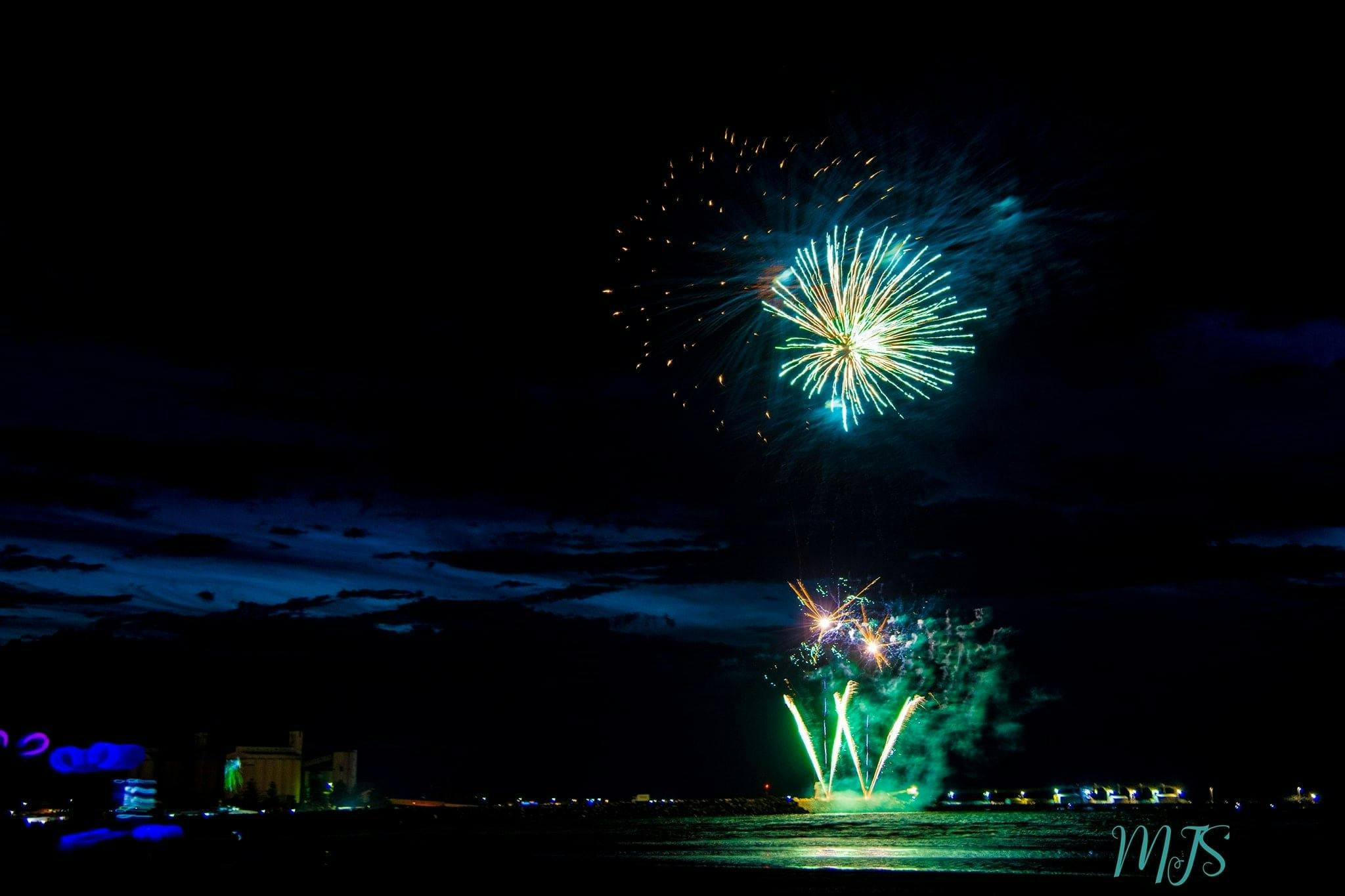 a speckled fire work disappearing as a aqua, golden fireworks explode over the night sky and sea.