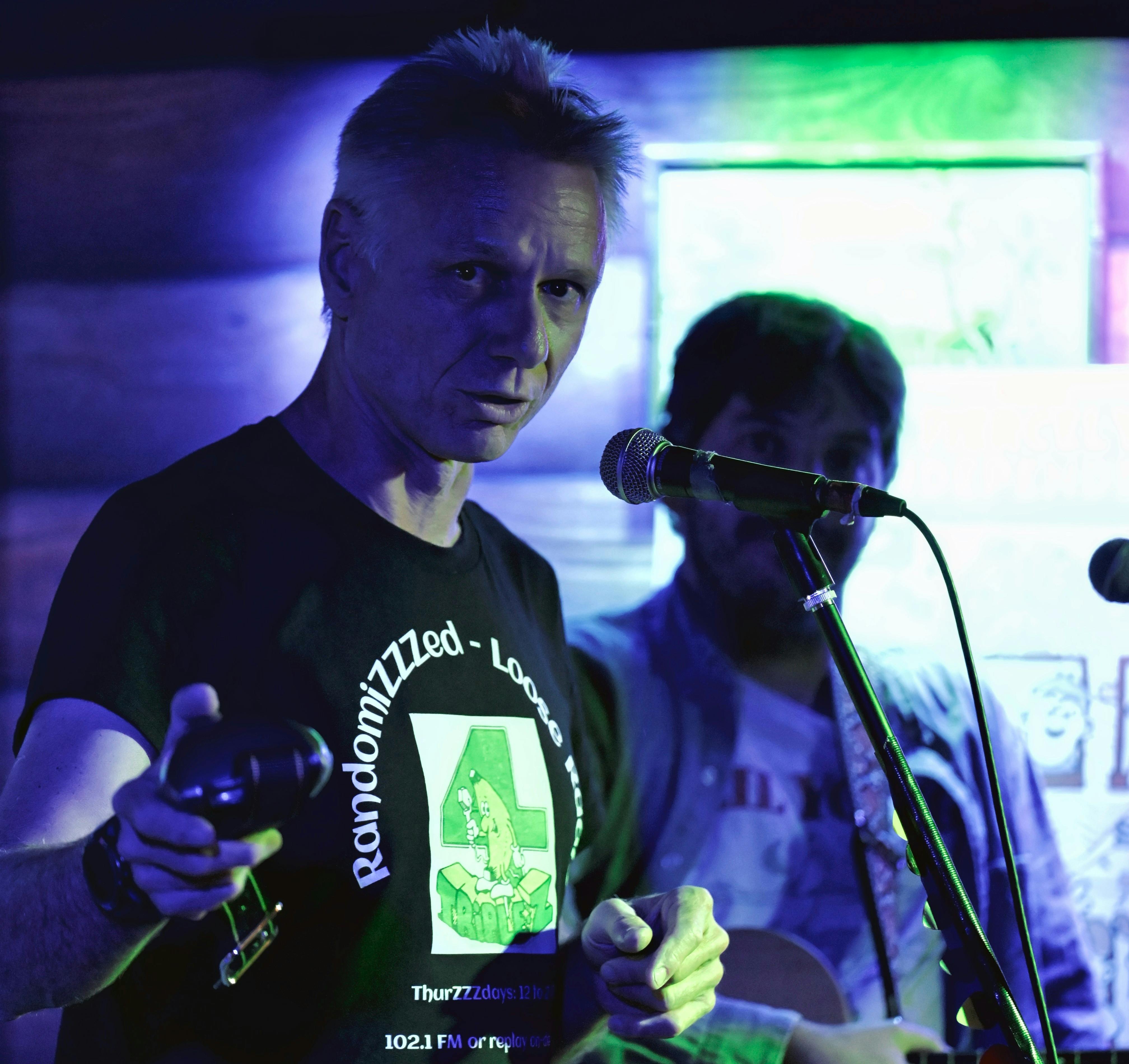 Two people on a stage, man on left has a black tee shirt with banana log and is holding a cowbell.