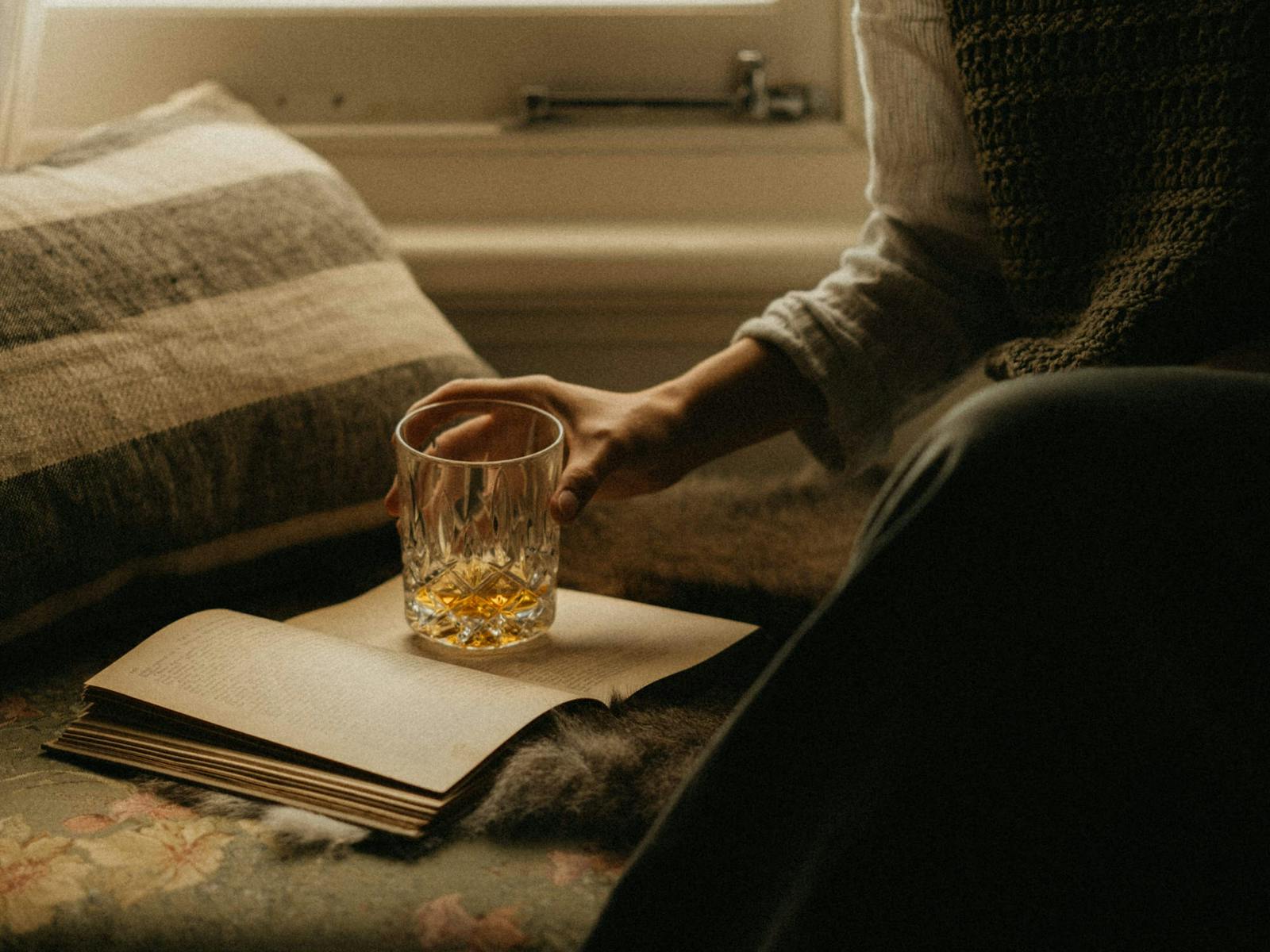 a woman's hand reading a book and holding a glass of whisky
