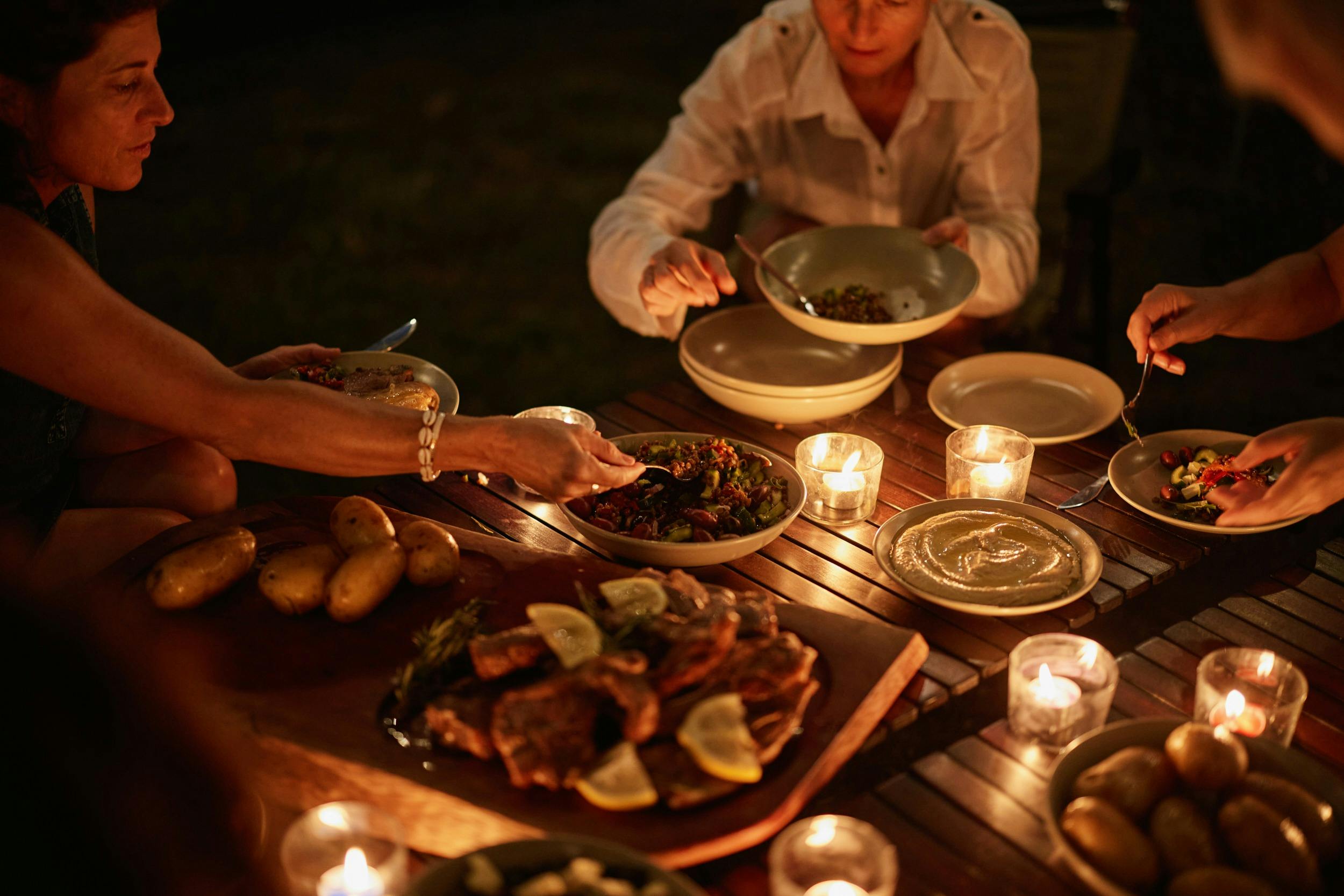 People serving themselves from a candlelit table spread with boards and bowls filled with food.