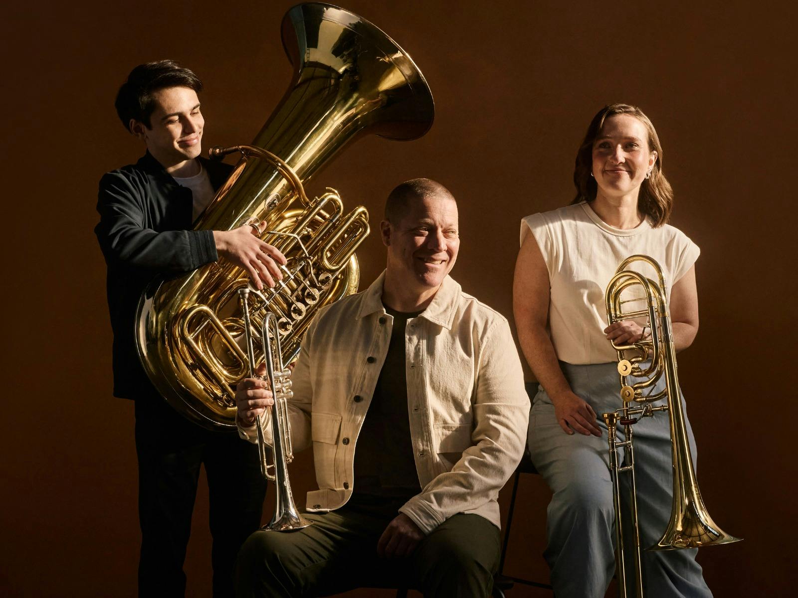 A photo of  three ASO brass players holding their instruments and smiling in a studio setting.