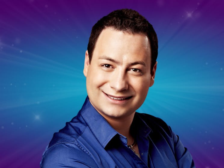 Man in blue button up shirt smiling in front of a blue sparkled backdrop.