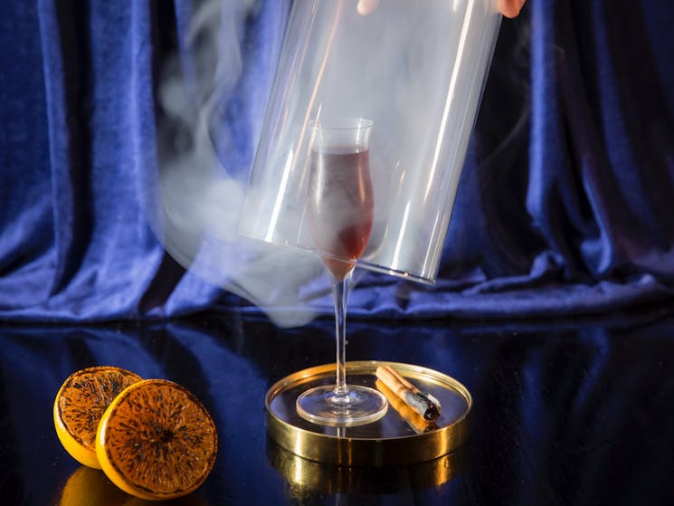 A cloche being lifted over a smoke filled glass with a cinannamon stick