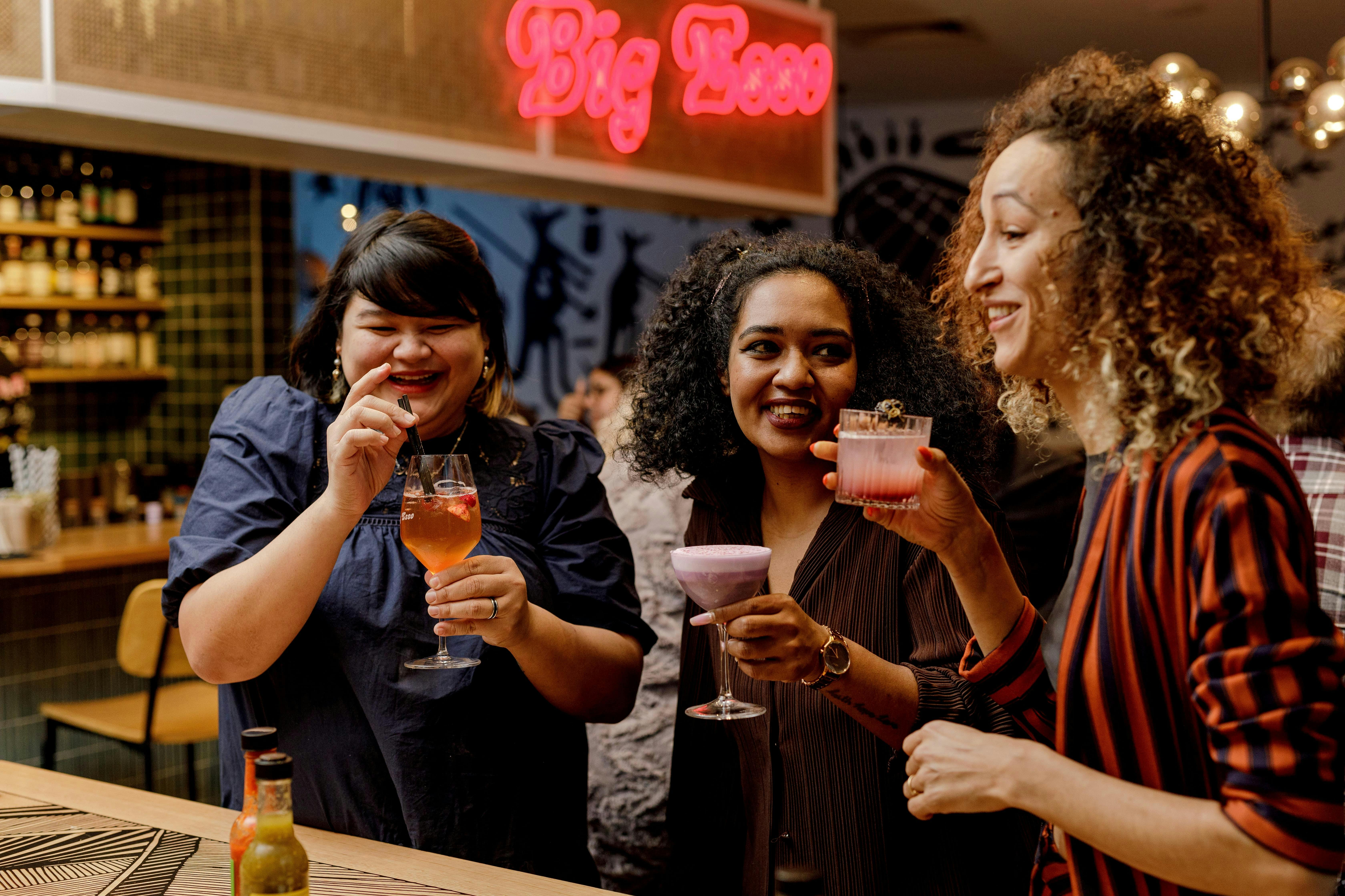 3 women standing at hi table sipping cocktails and laughing.