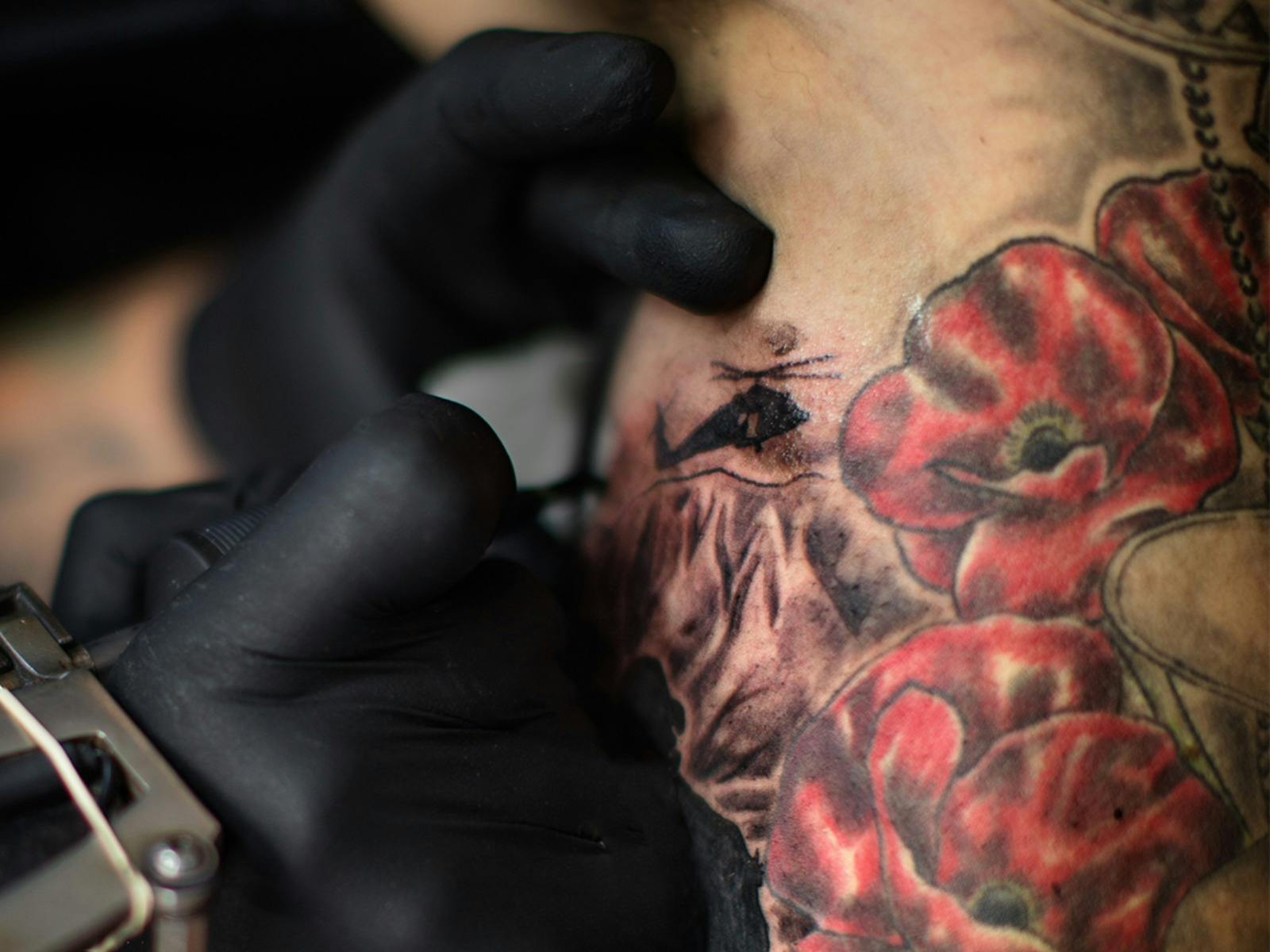 Close-up of a gloved tattoo artist working on skin next to red poppy tattoos.