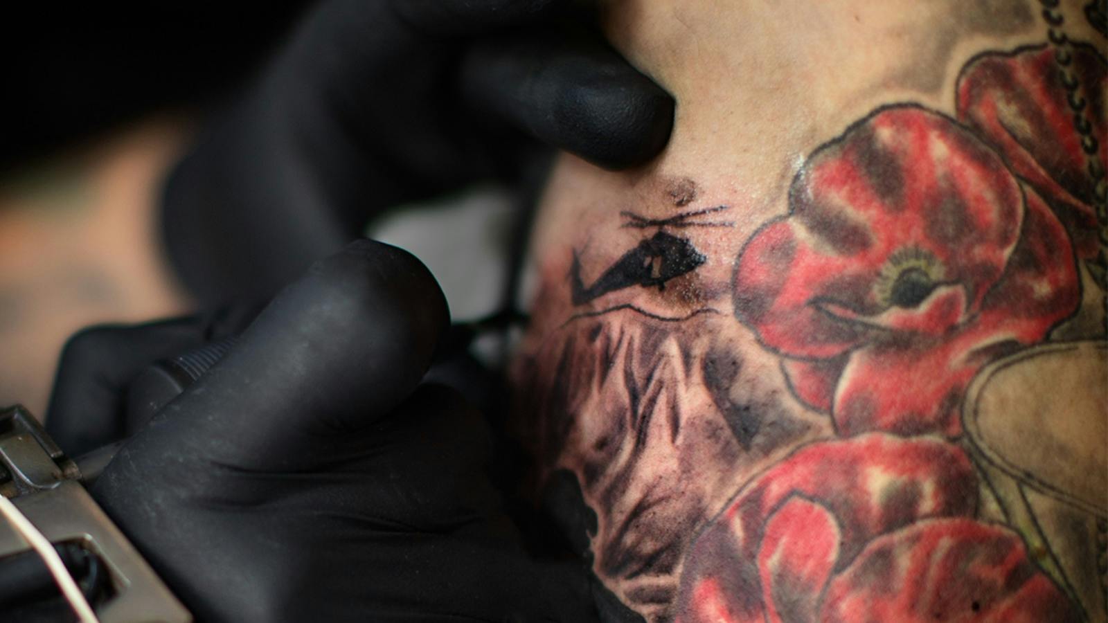 Close-up of a gloved tattoo artist working on skin next to red poppy tattoos.