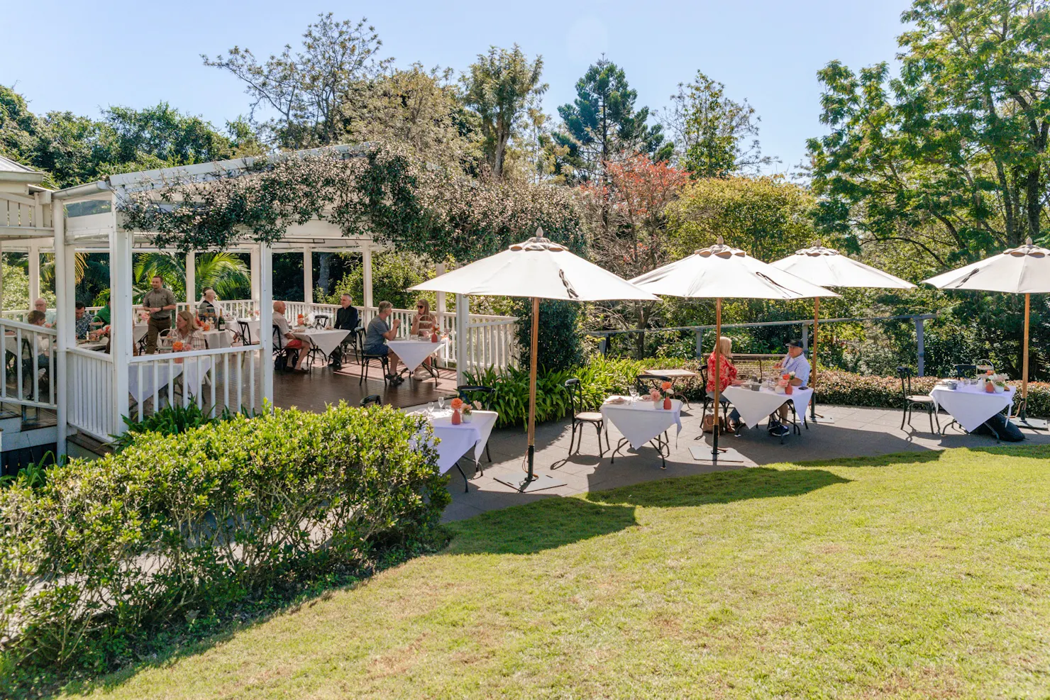 People dining at an outdoor patio and tables with large umbrellas by the gardens