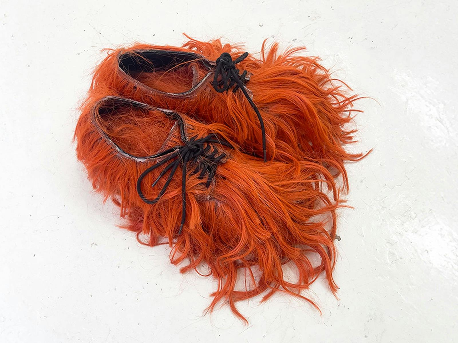 A pair of shoes covered in long, vivid orange synthetic hair, with black laces still visible.