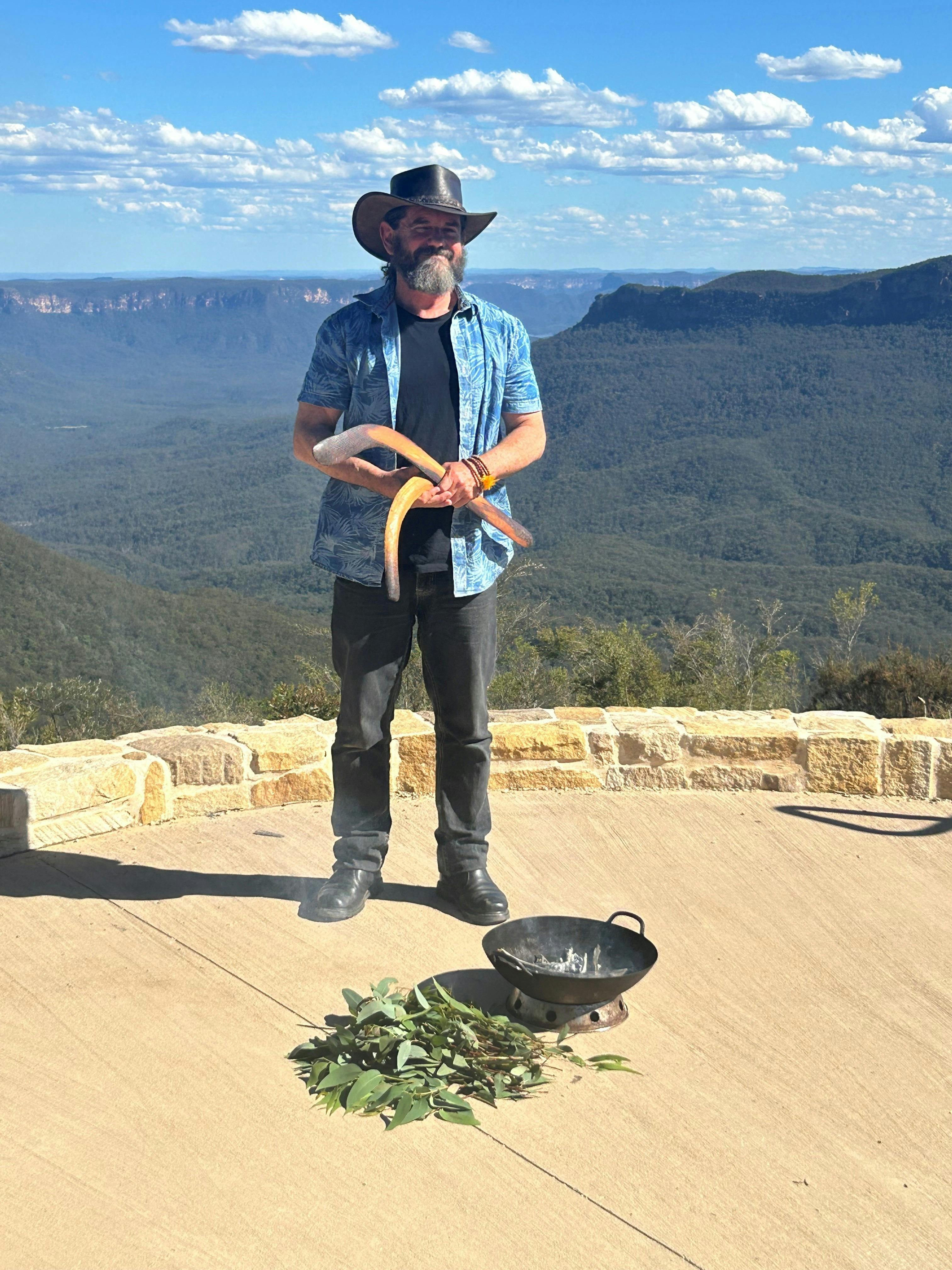 A man in a hat holding clap sticks stands in front of a landscape view