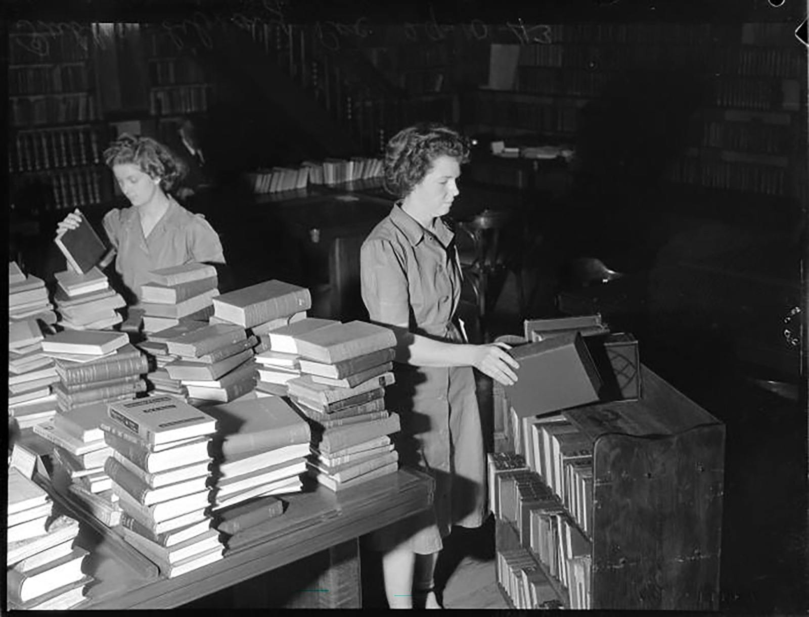 A black and white photo of two women working in a library amongst stacks of books