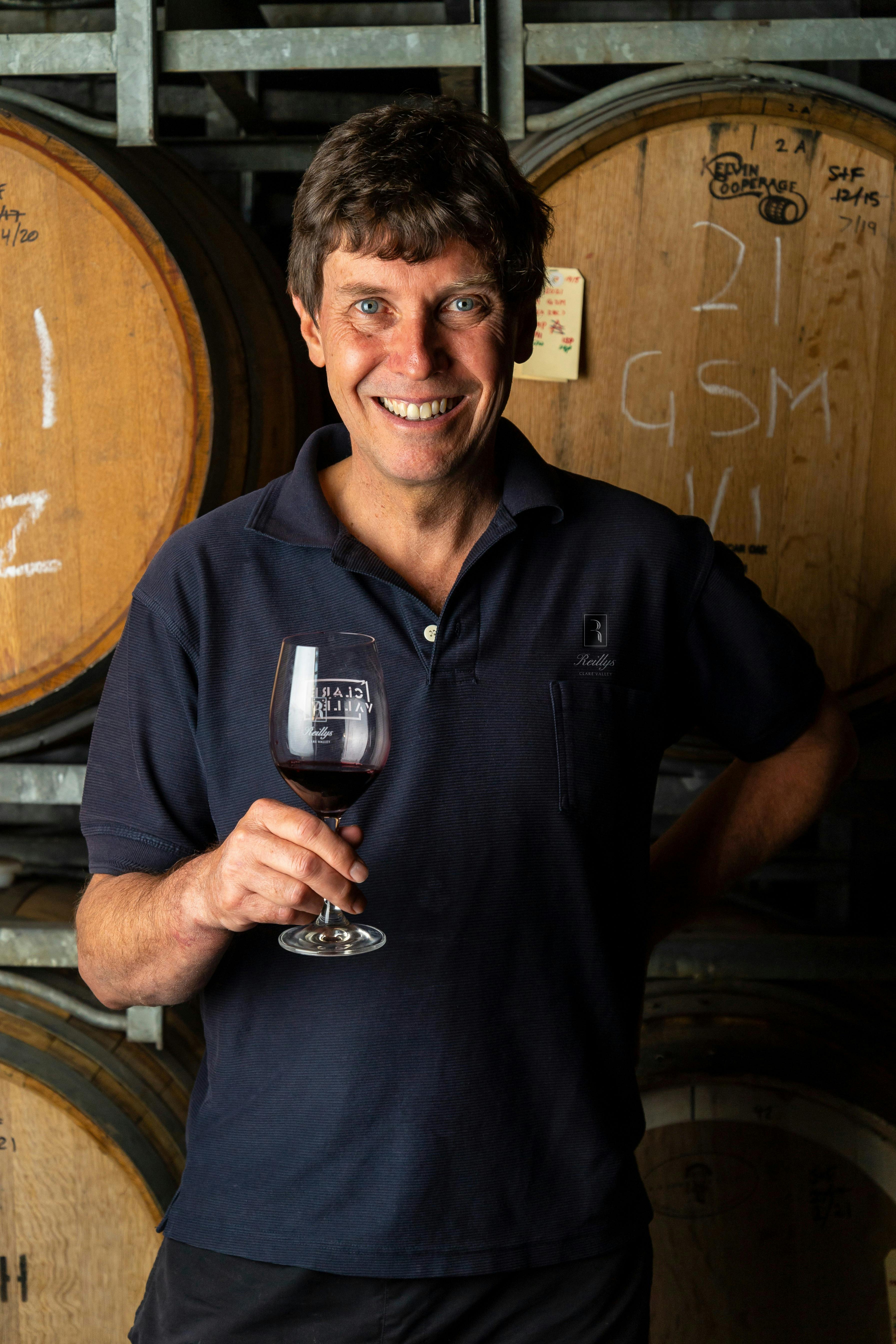 Reillys' winemaker Justin standing in front of wine barrels smiling holding glass of red wine
