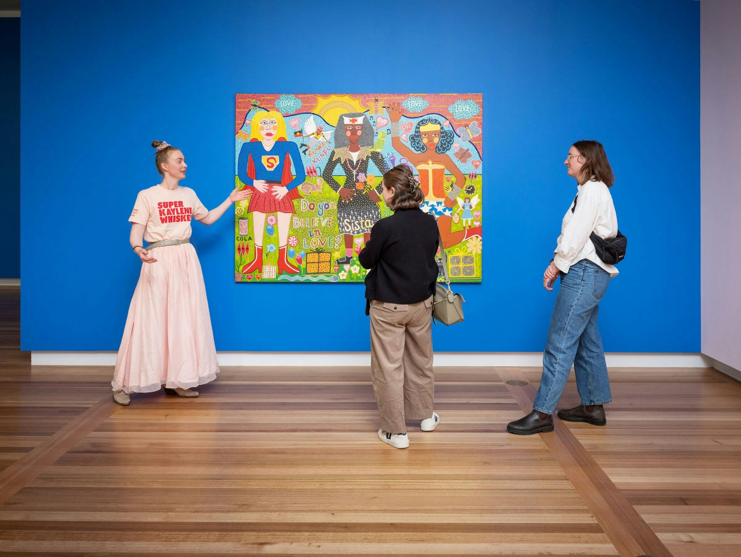A guide in a pink outfit speaks to two visitors about a colourful portrait.