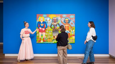 A guide in a pink outfit speaks to two visitors about a colourful portrait.