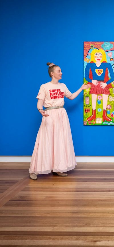 A guide in a pink outfit speaks to two visitors about a colourful portrait.