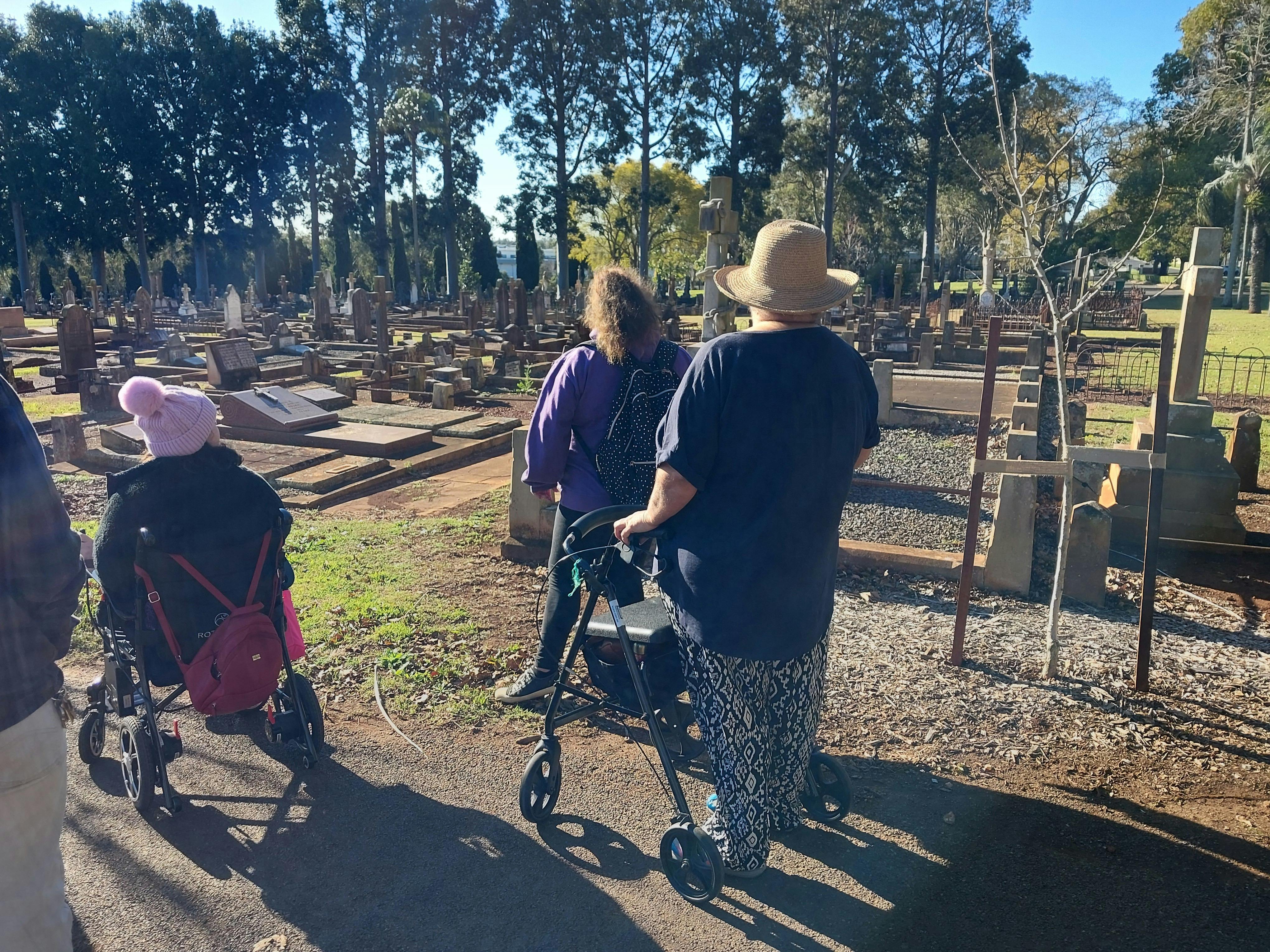 Several people, backs to camera, one using a wheelie-walker, one in a wheelchair, looking at graves.