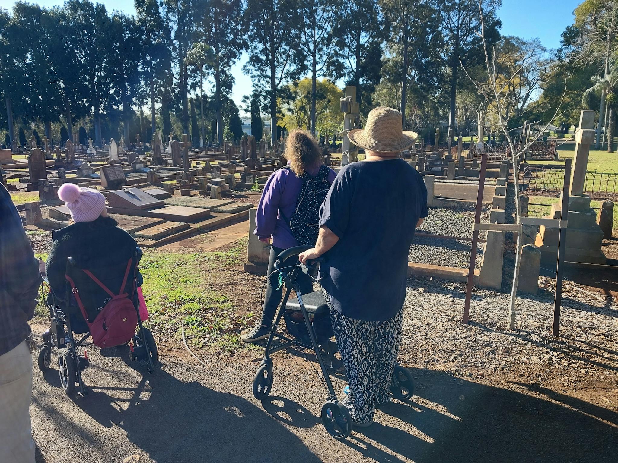 Several people, backs to camera, one using a wheelie-walker, one in a wheelchair, looking at graves.