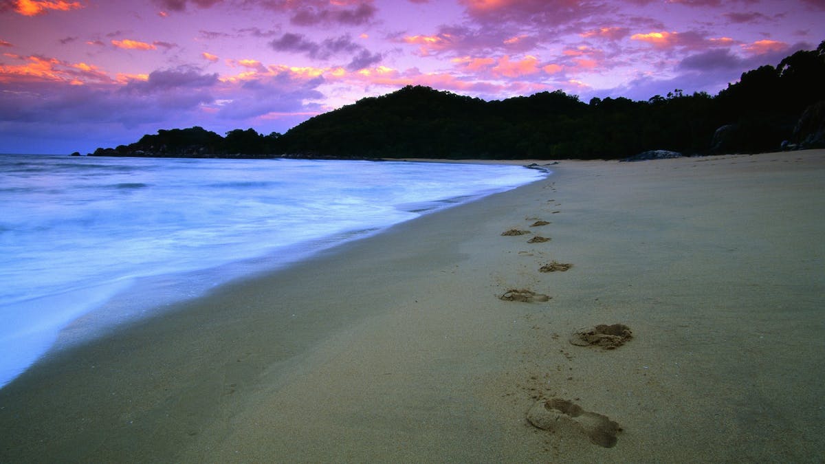 Water laps against the beach with distant headland silhouetted under pink sunset skies. Water laps against the beach with distant headland silhouetted under pink sunset skies.