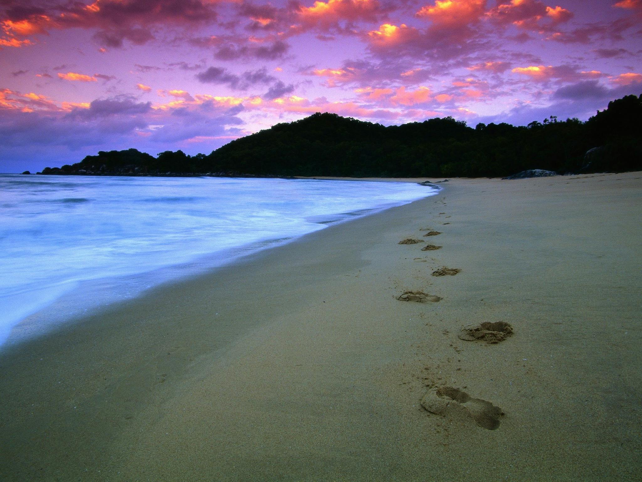Water laps against the beach with distant headland silhouetted under pink sunset skies.