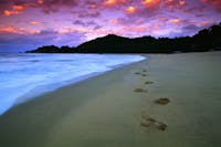 Water laps against the beach with distant headland silhouetted under pink sunset skies.