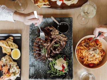 Two hands holding forks over a T-bone steak and spaghetti, representing a main meal with a bonus sid