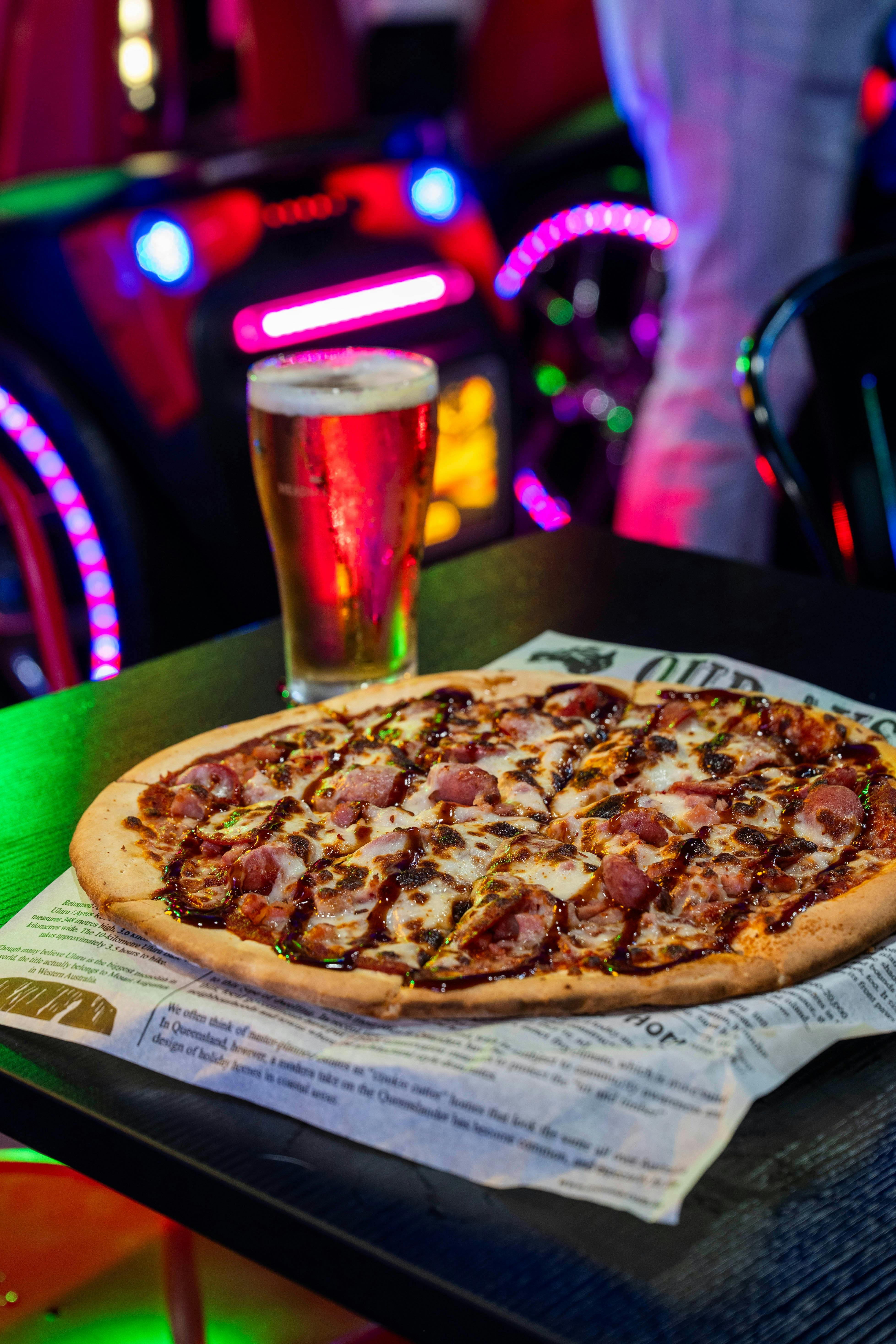 Closed-up of a meat lovers  pizza and a beer on a table with neon arcade lights in the background