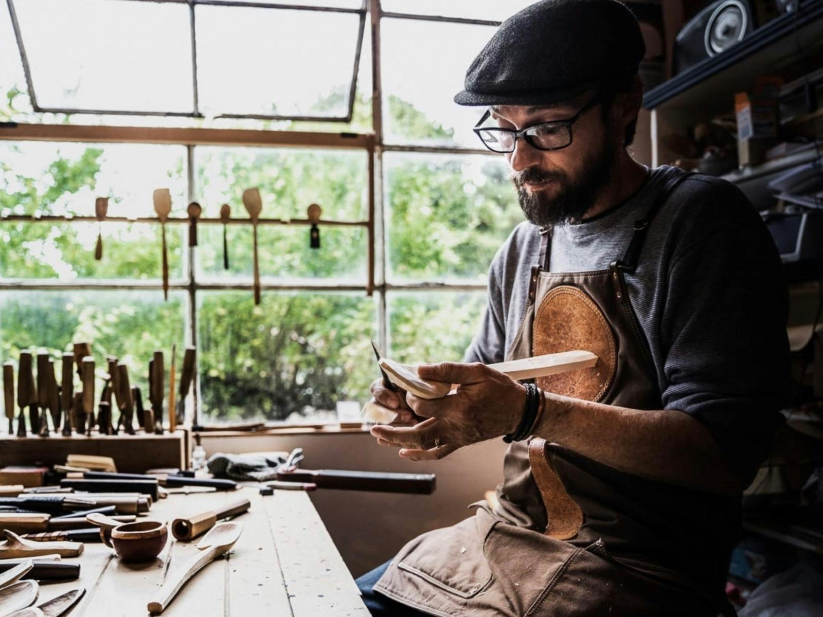 A spoon carver in leather apron sits by a sunlit window carving wooden spoons