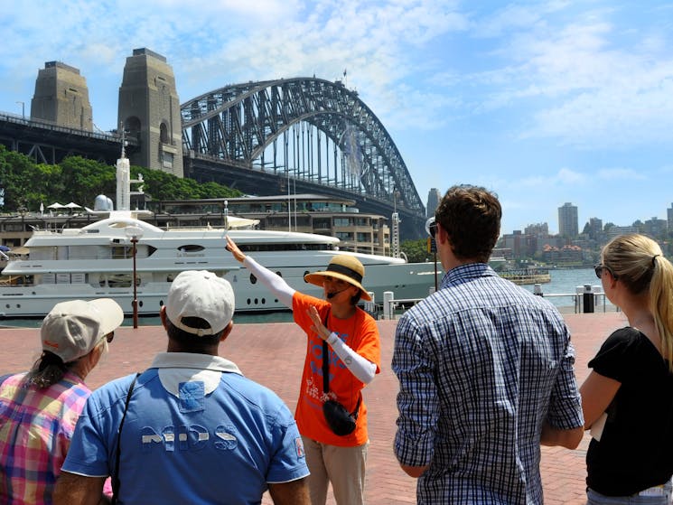 Free walking tour Sydney is at the Rocks. A tour guide in orange uniform uncovers local history.