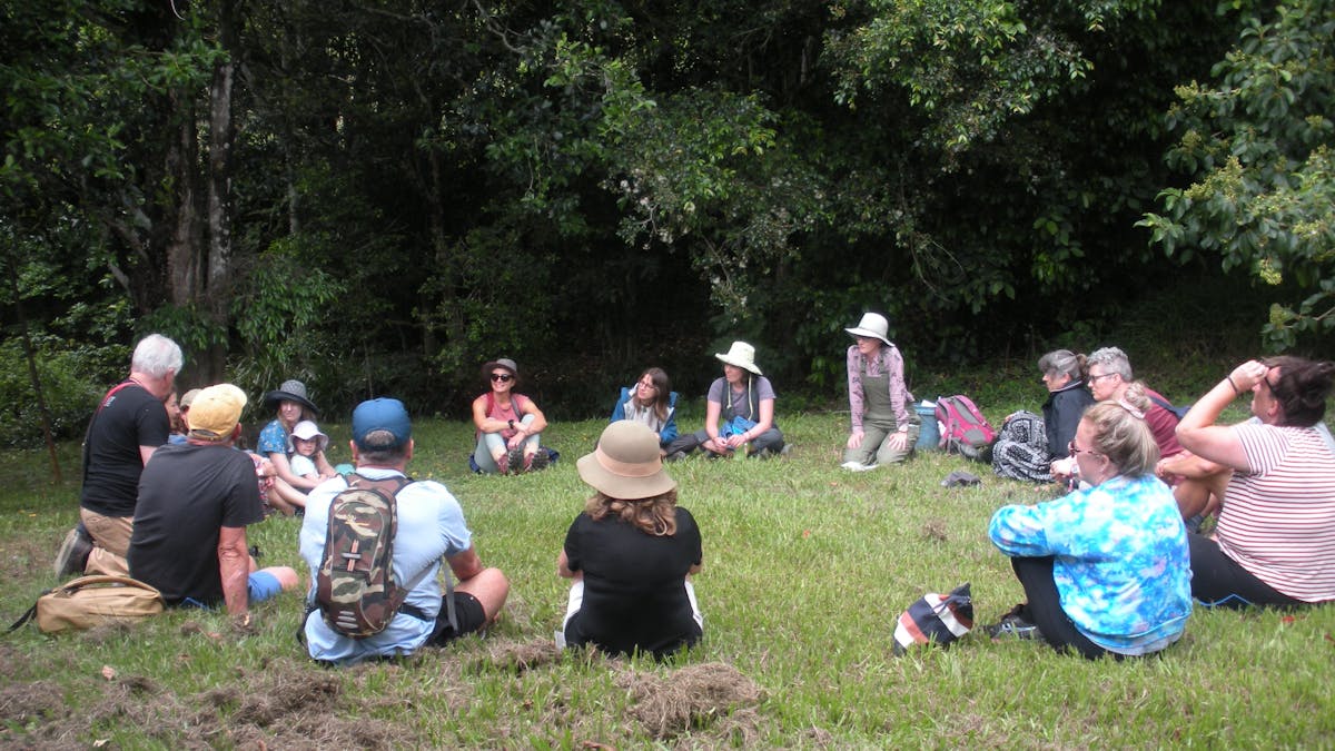 group of adults and children sitting on grass under trees with hats and backpacks