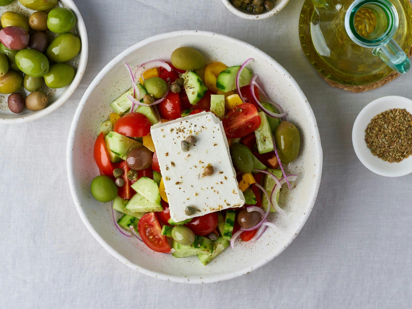Overhead view showing serving bowl of salad with wedge of feta on top. Bowl of olives to the left.