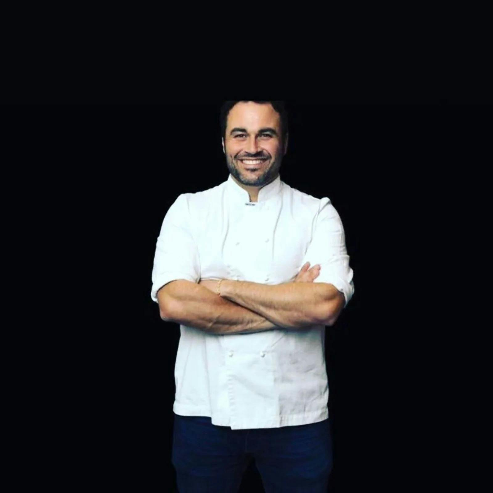 Chef, Miguel Maestre, wearing a white chef’s jacket, standing against a dark background