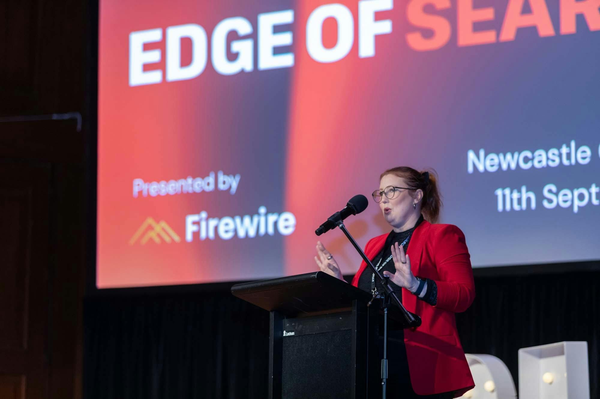 Woman in red jacket speaking at a lecturn in front of a screen that says Edge of Search.