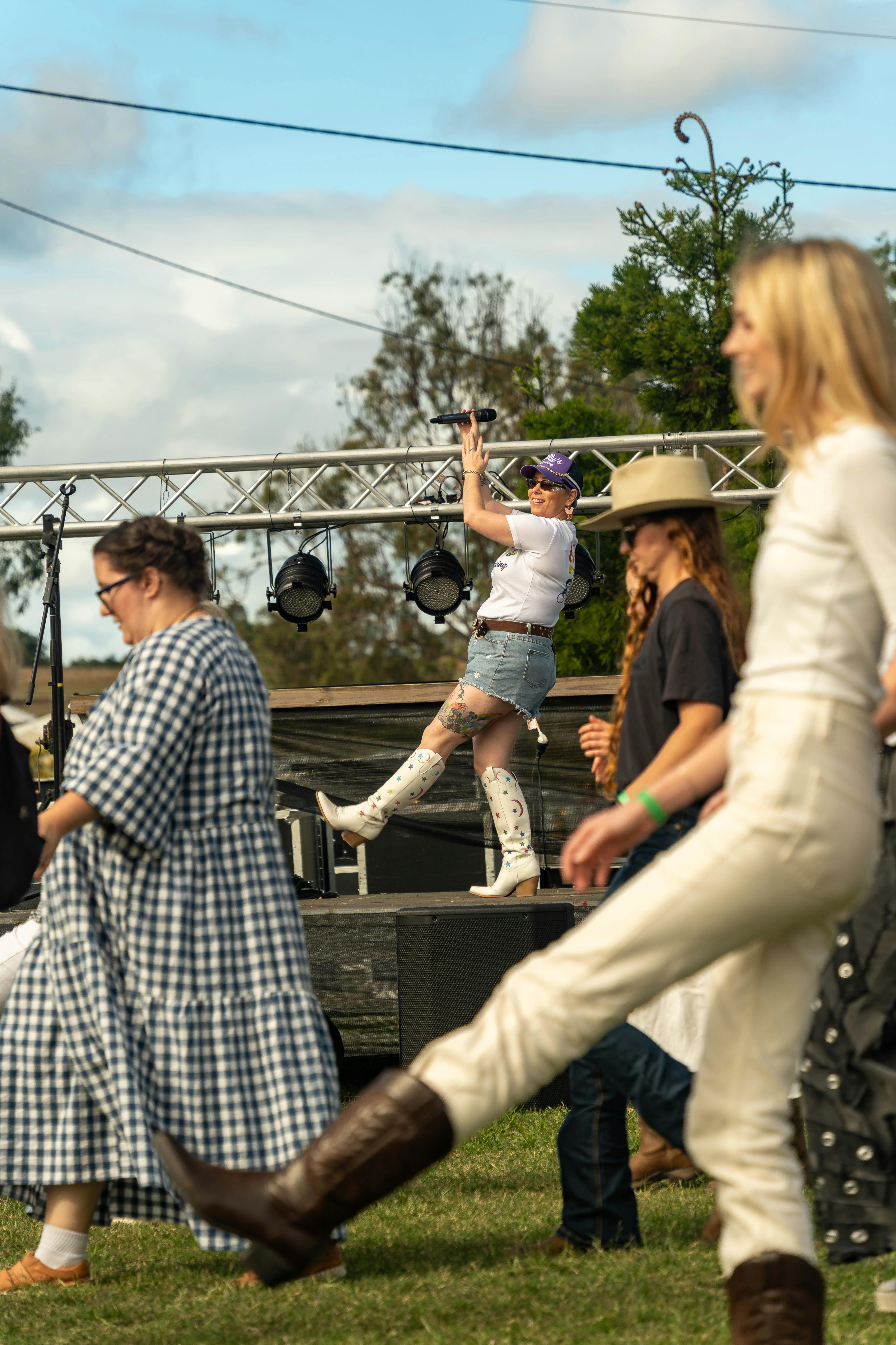A woman on stage teaching the crowd line dancing.  You can see the dancers on the grass dancing