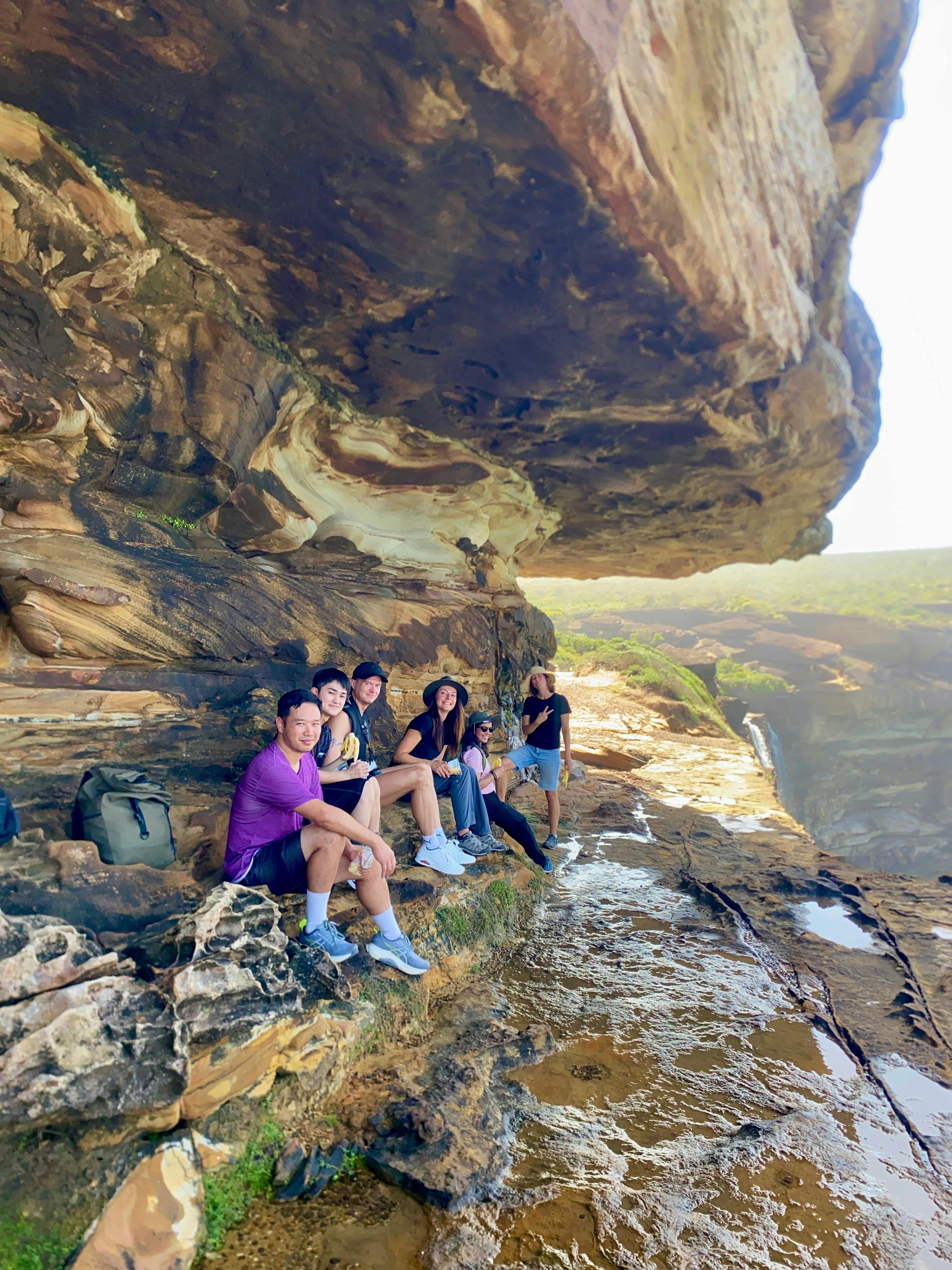 Group of hikers resting under a large sandstone overhang with sea views behind