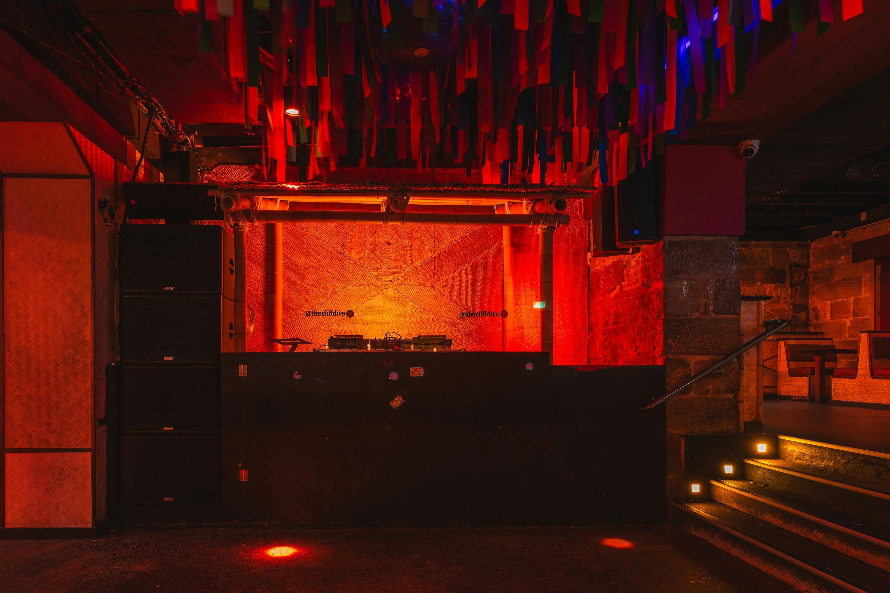 A nightclub DJ booth lit in deep red and orange light, framed by bamboo accents and stone walls