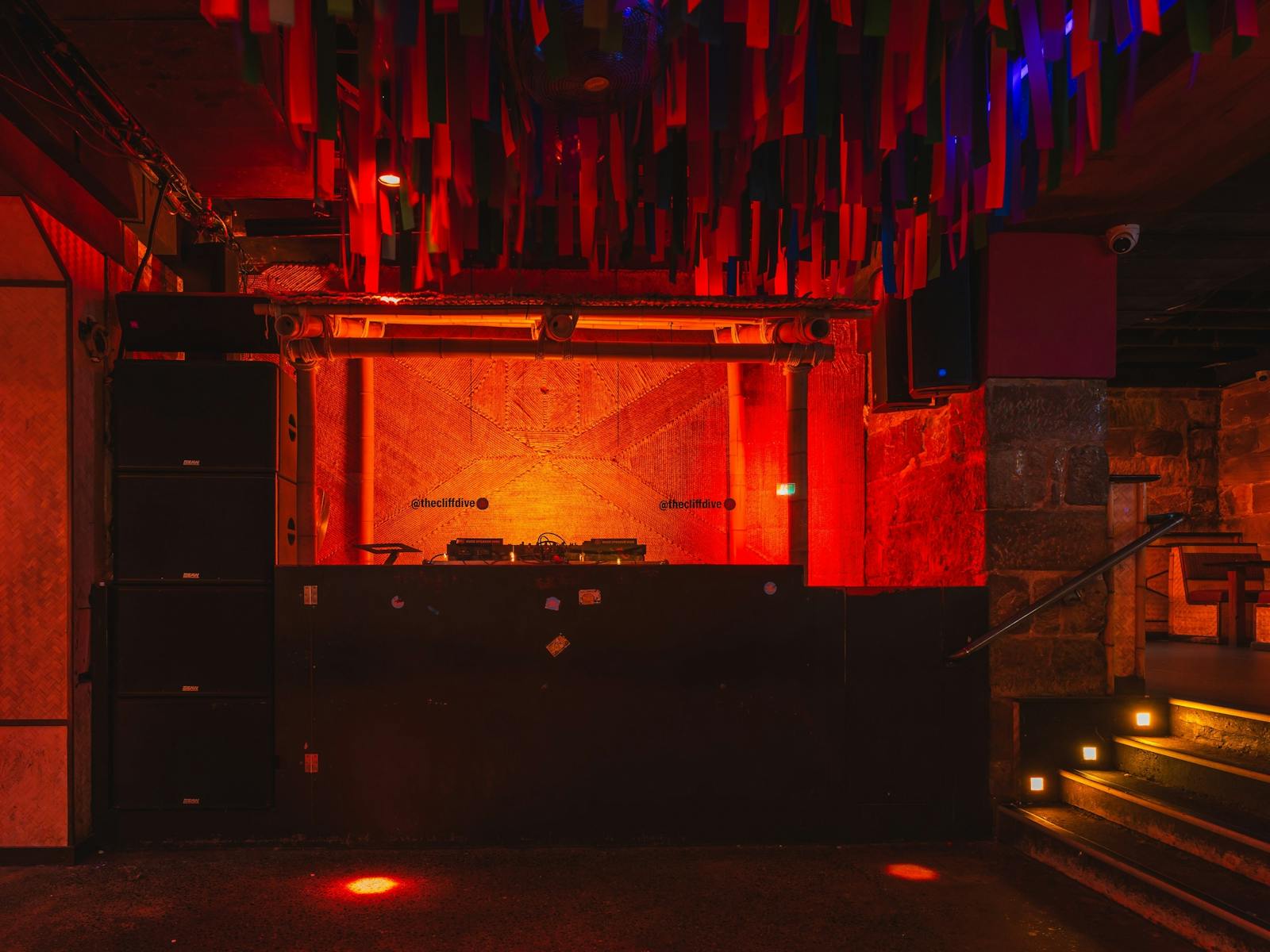 A nightclub DJ booth lit in deep red and orange light, framed by bamboo accents and stone walls