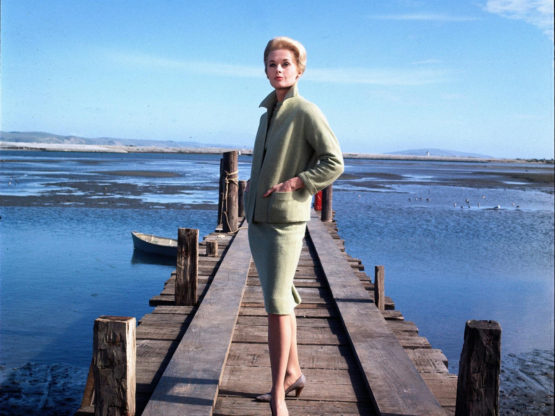 Woman dressed in suit stands on pier looking out past the camera, water in background