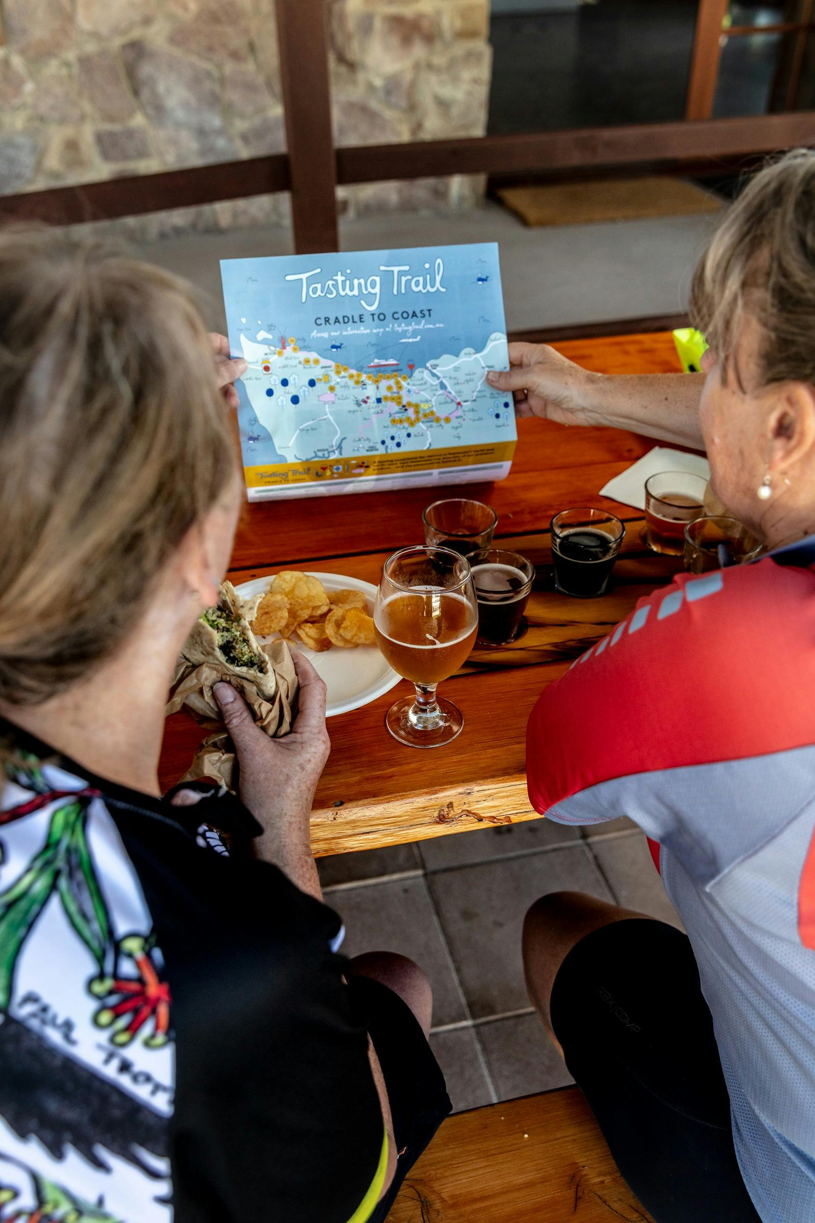 Two women looking at a Tasting Trail map while eating wraps and drinking beer.