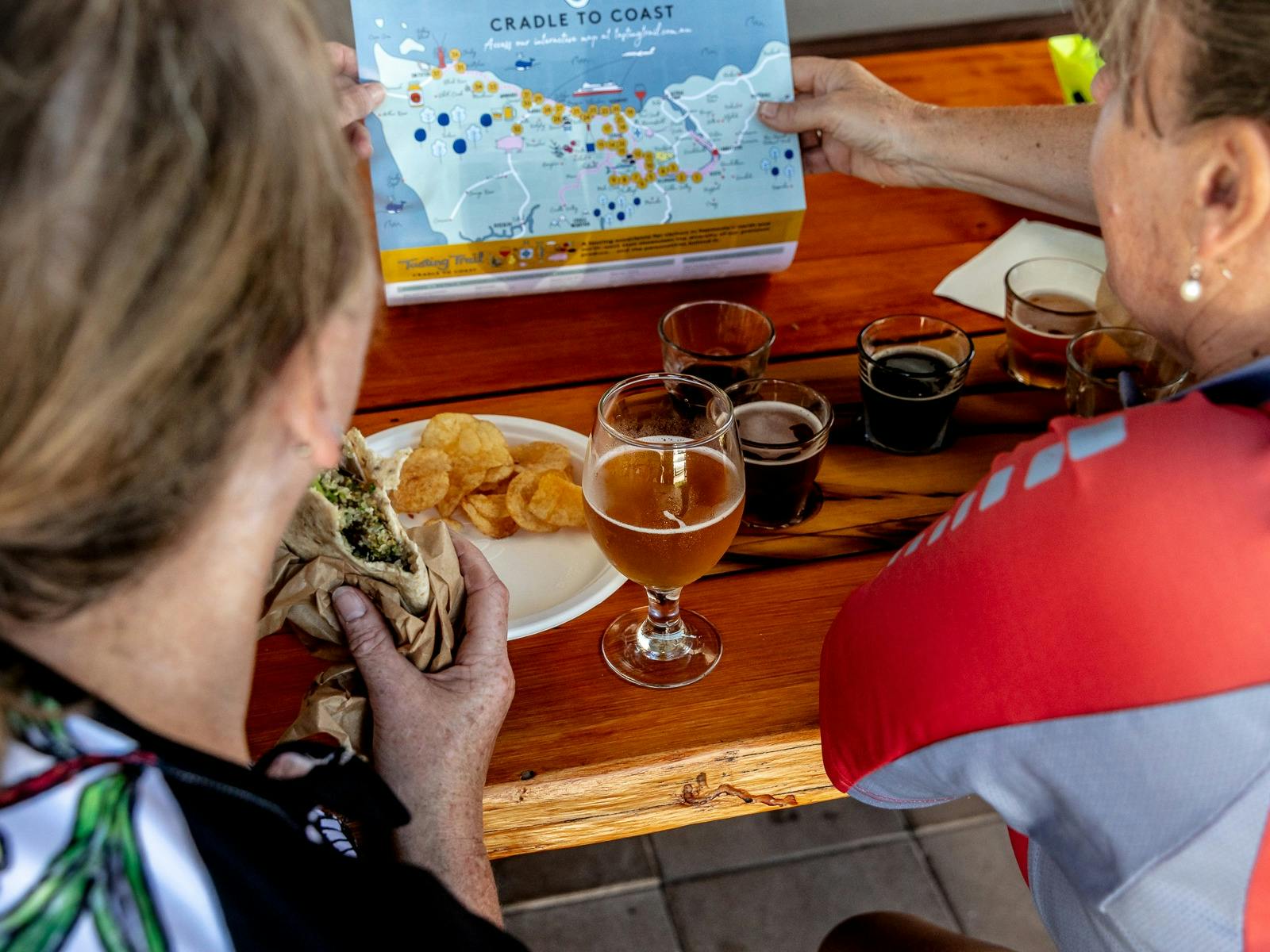 Two women looking at a Tasting Trail map while eating wraps and drinking beer.